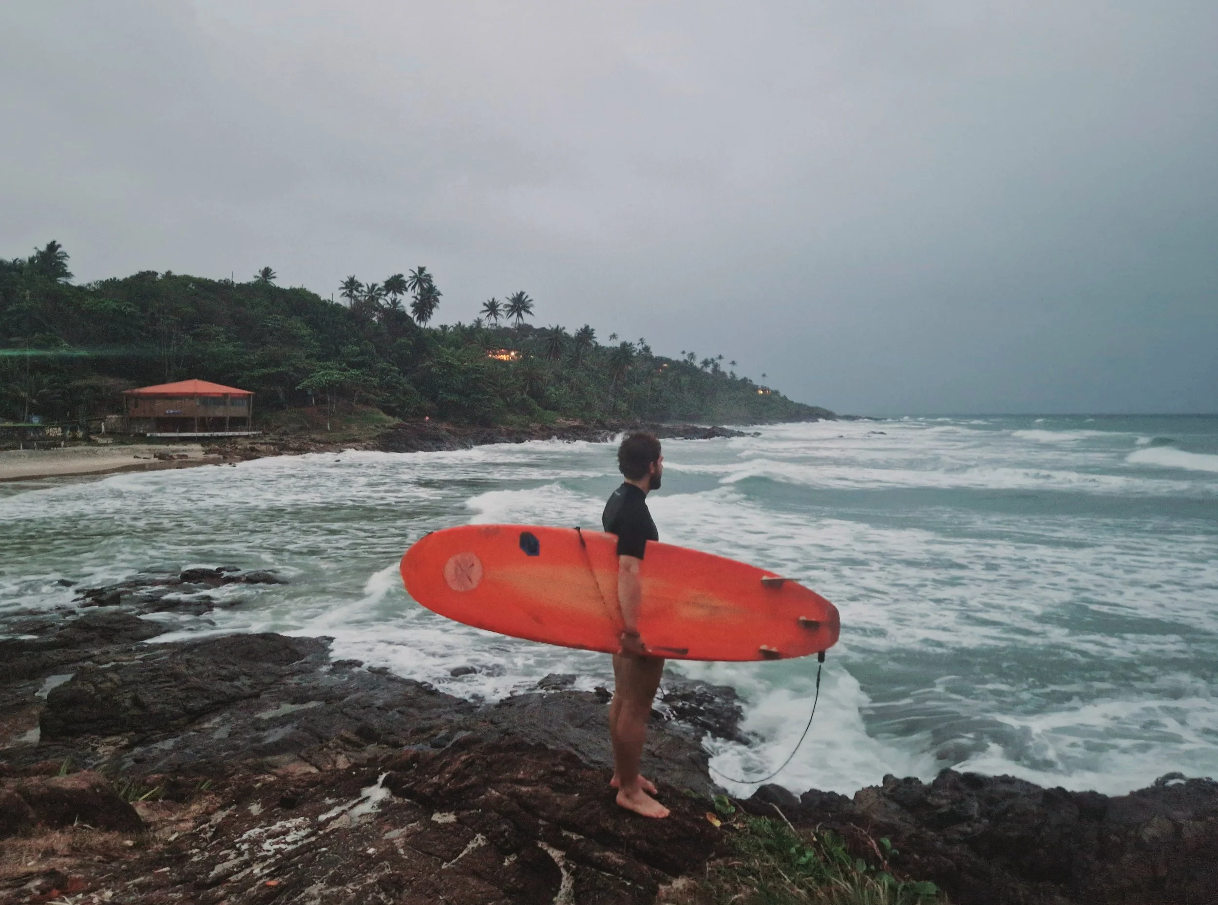 A man holding an orange surfboard stands on rocky shore facing the ocean, with waves and a coastal landscape with trees and buildings in the background.