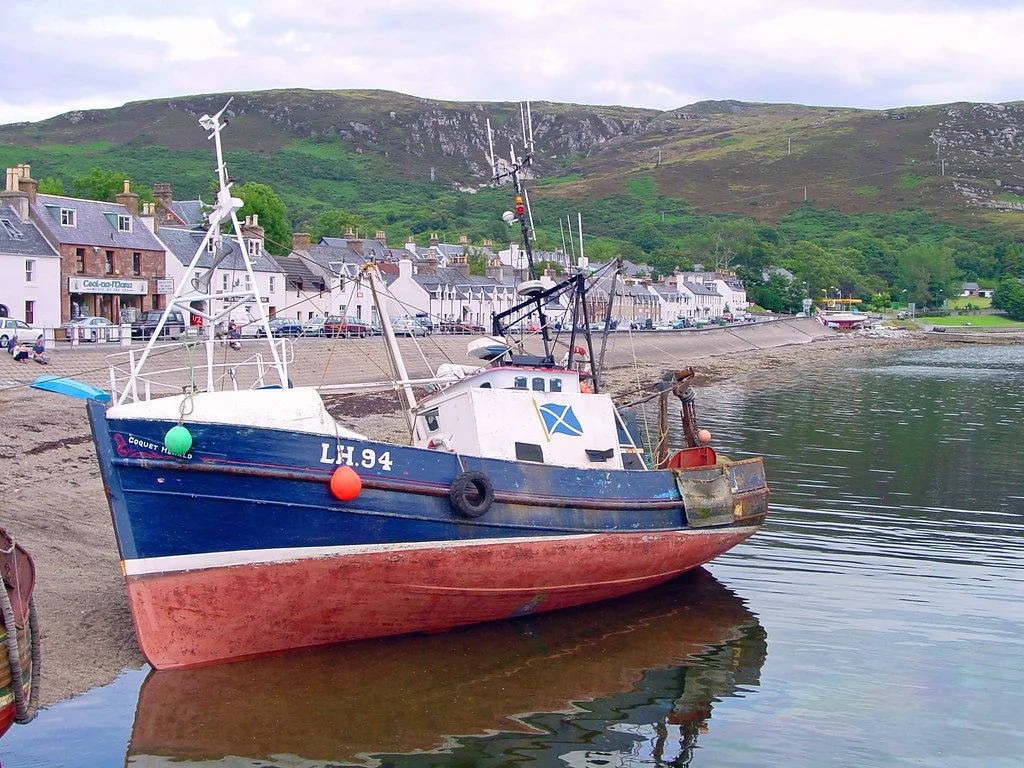 A fishing boat with the registration LH.94 is docked on a pebble beach in a small Scottish coastal town, with white houses and green hills in the background.