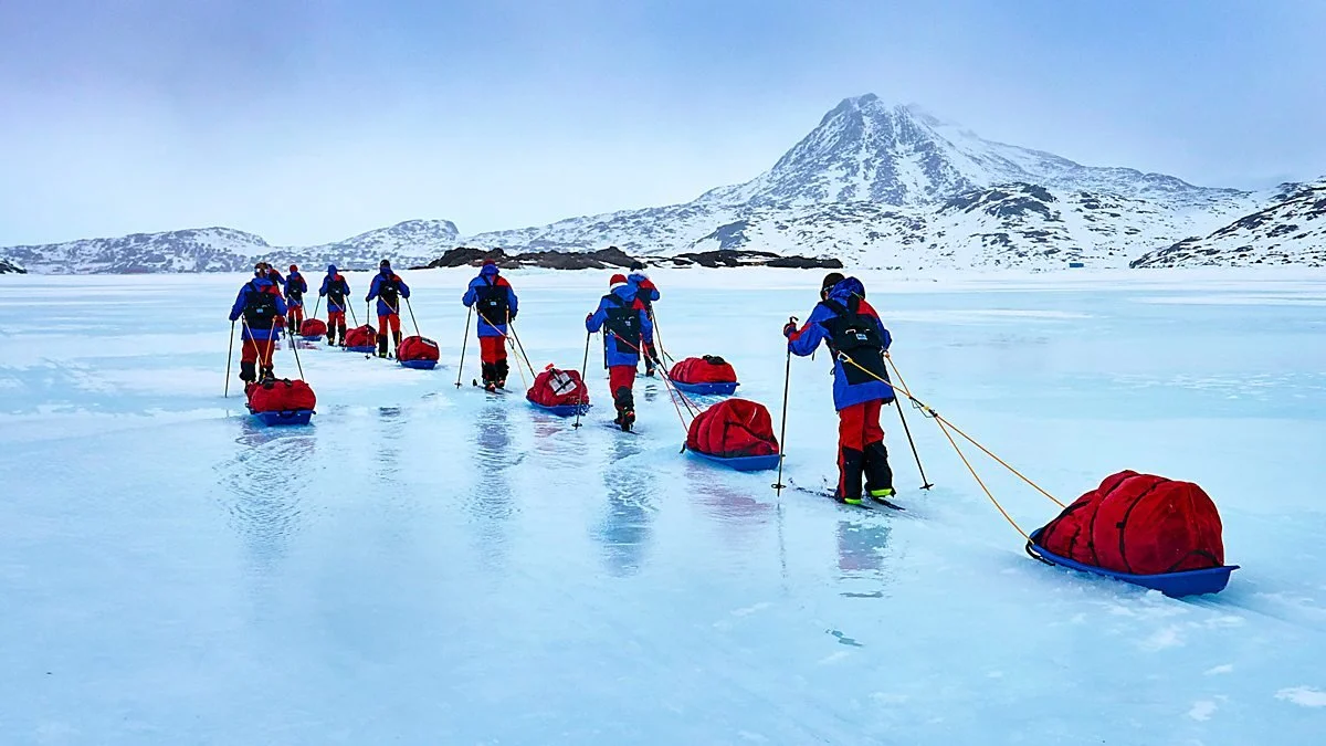 Group of people walking across a frozen landscape with snow-covered mountains in the background, pulling sleds loaded with gear.