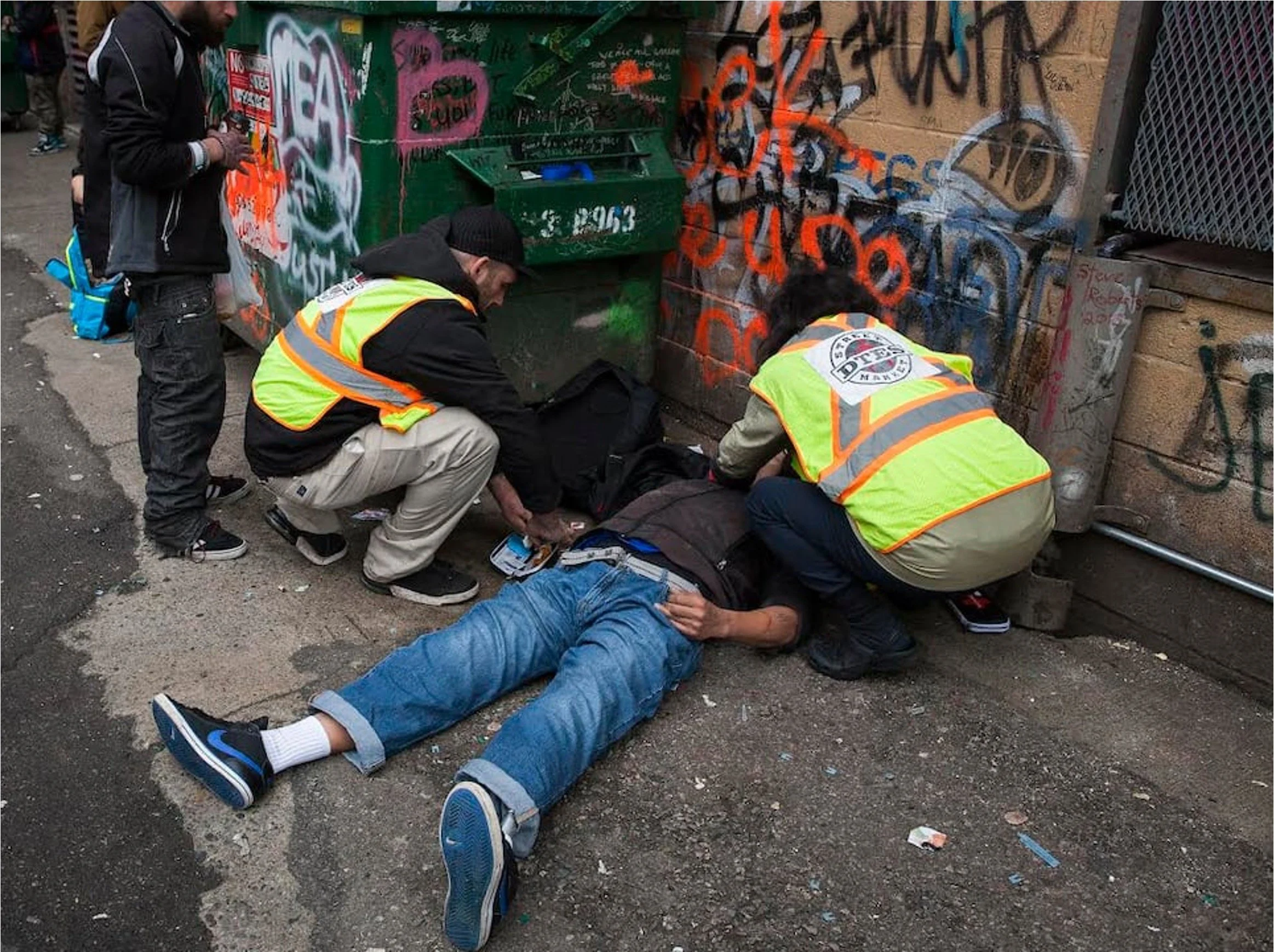 Three people assisting an unconscious person lying on the ground next to a graffiti-covered wall and green dumpster.