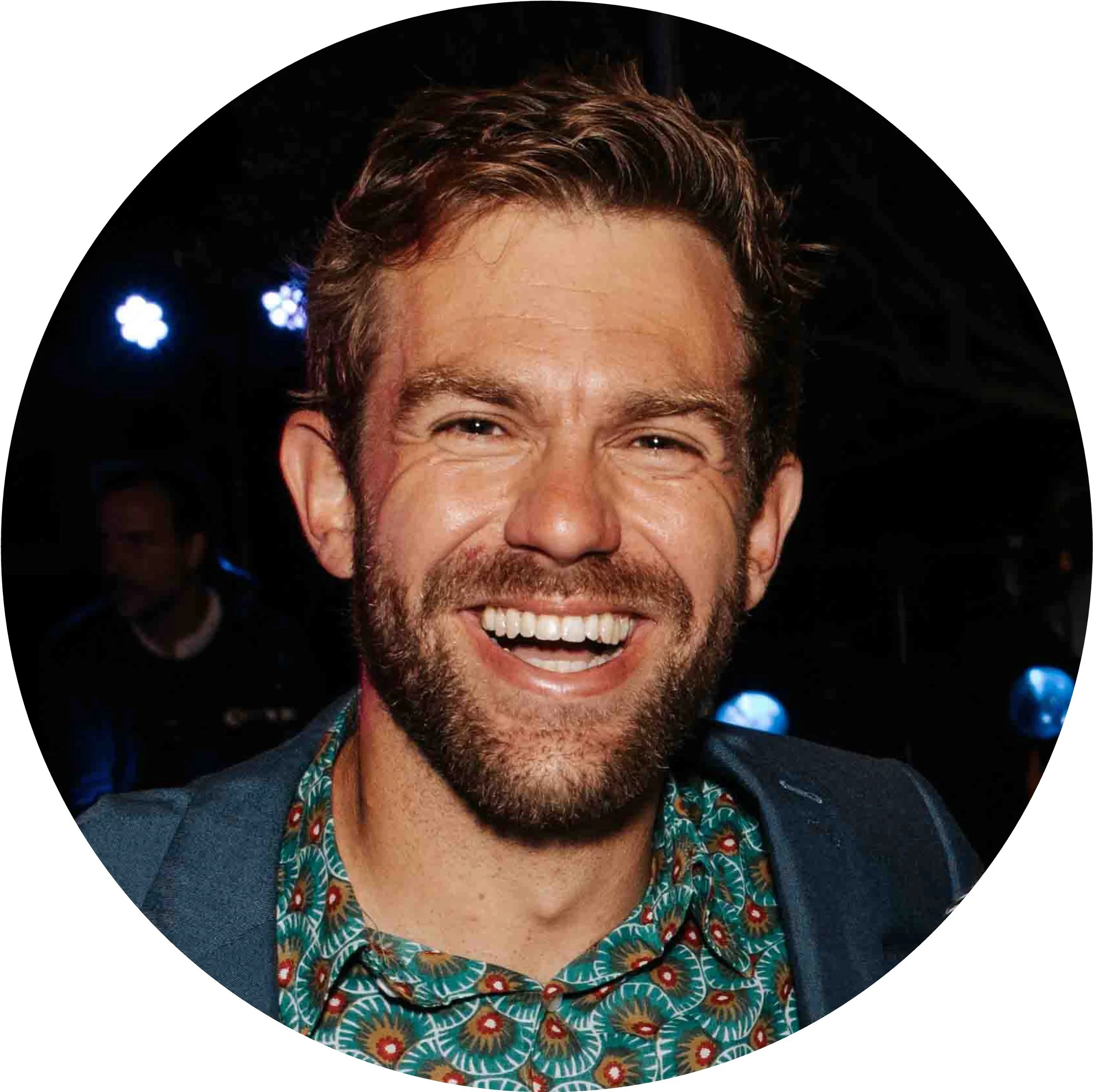 A smiling man with a beard and brown wavy hair, wearing a patterned shirt and a blazer, in a setting with dark background and blue lights.