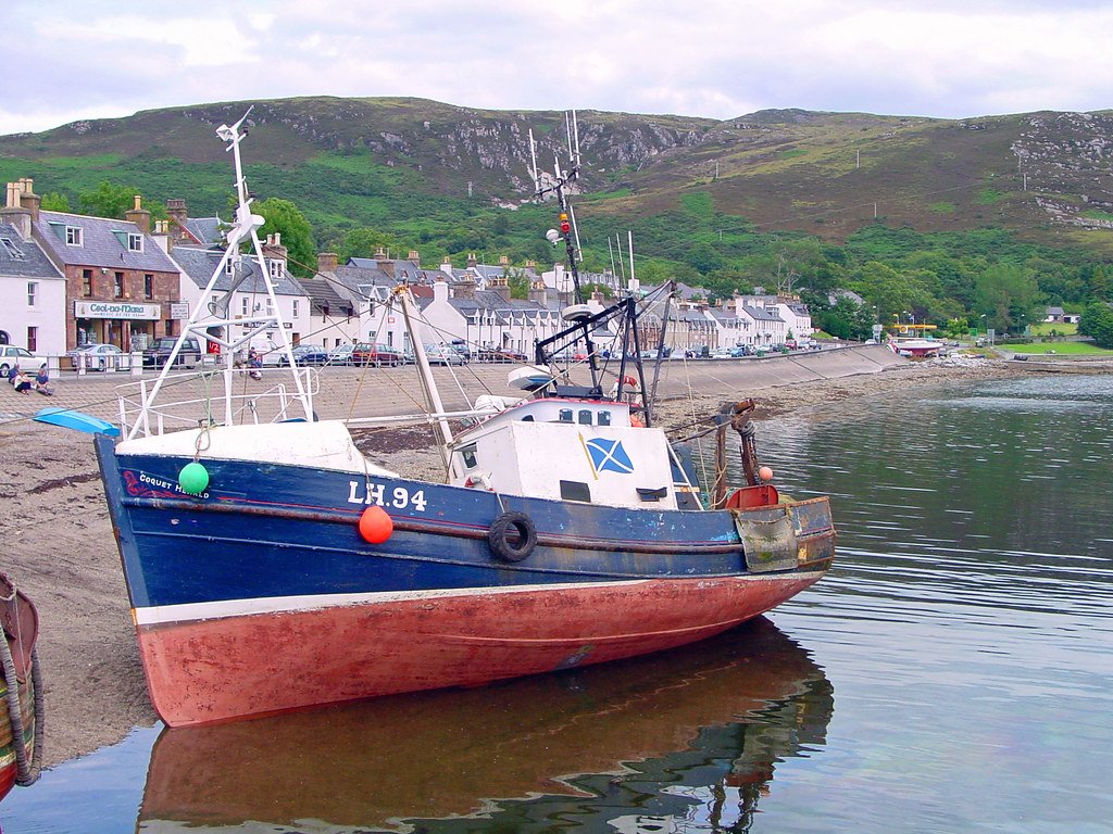 Fishing boat moored near a small coastal village with white houses, green hills, and a cloudy sky.