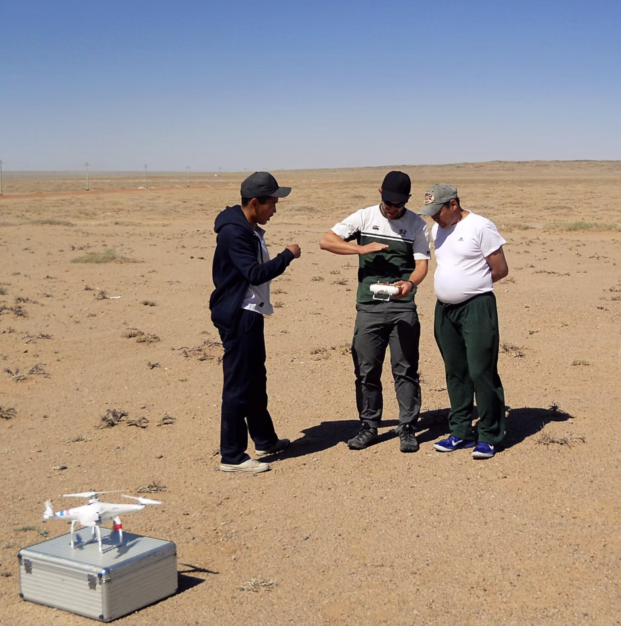 Three men standing in a desert, with a drone resting on a nearby metal case. Two men are looking at a remote control device, with one holding a phone. The men appear to be preparing or inspecting the drone.