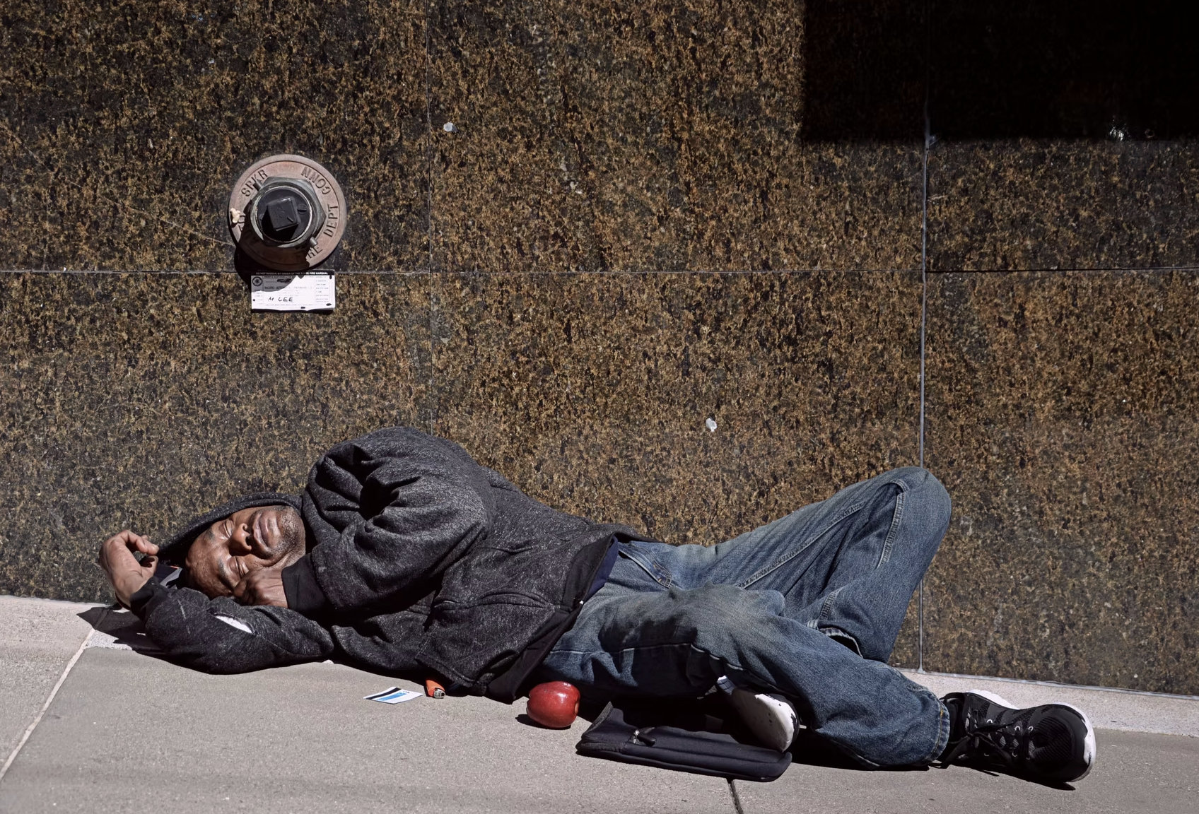 A man sleeping on the sidewalk, lying on his side with his head resting on his arm, against a brown tiled wall. He is wearing a dark jacket, jeans, and sneakers. Near his head, there is a black bag, a red apple, a paper or card, and a small object.