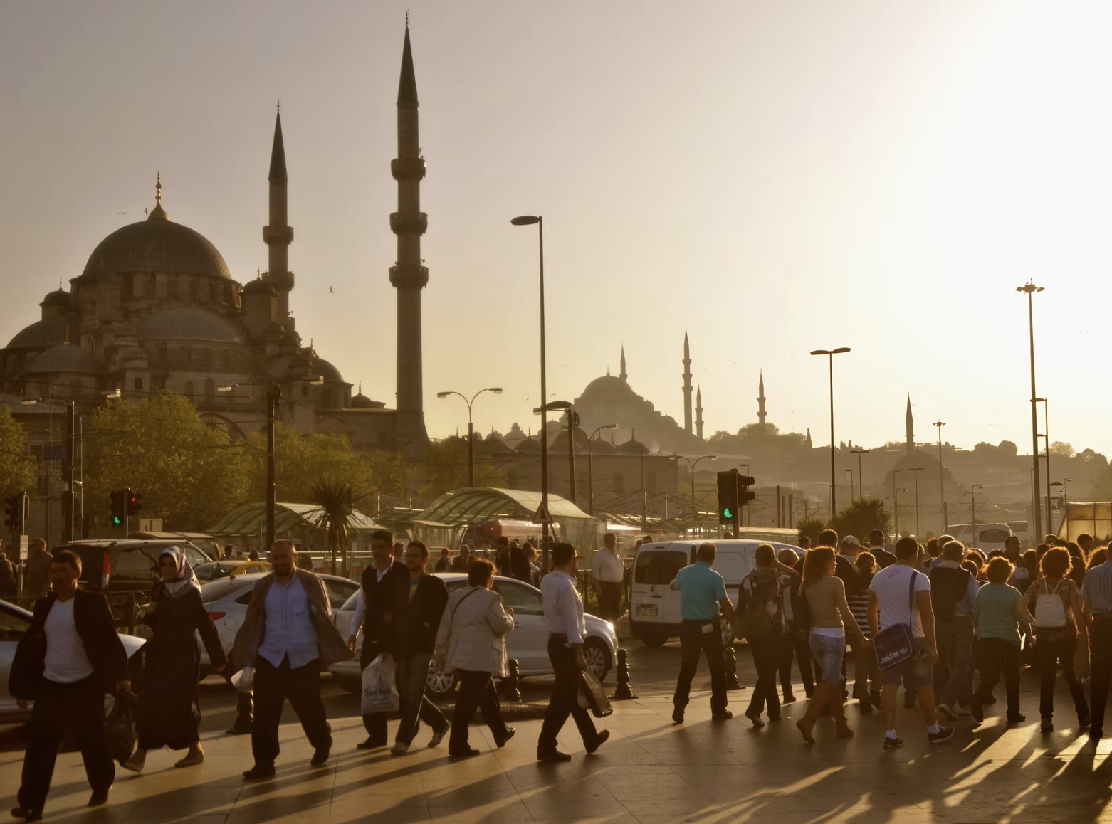 People walking on a busy city street at sunset with a mosque featuring domes and minarets in the background.