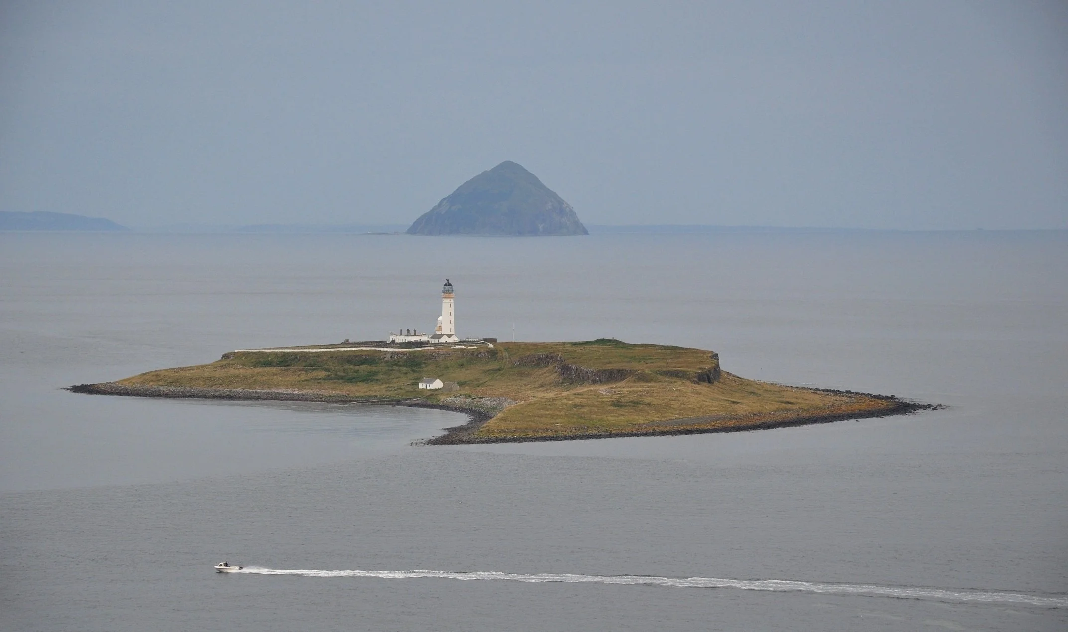 A lighthouse on a small island in a large body of water with a boat passing by, and a mountain in the background.