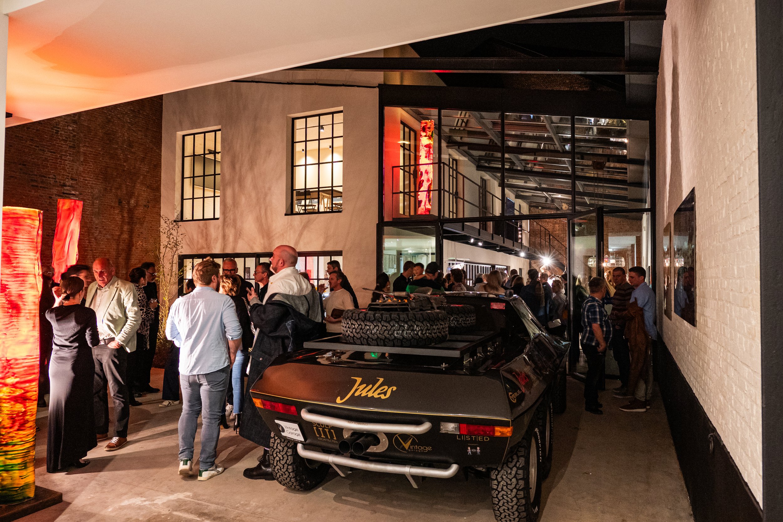 People gathered indoors at a social event with a vintage race car labeled 'Jules' parked nearby, and large orange and red art pieces on the left side.