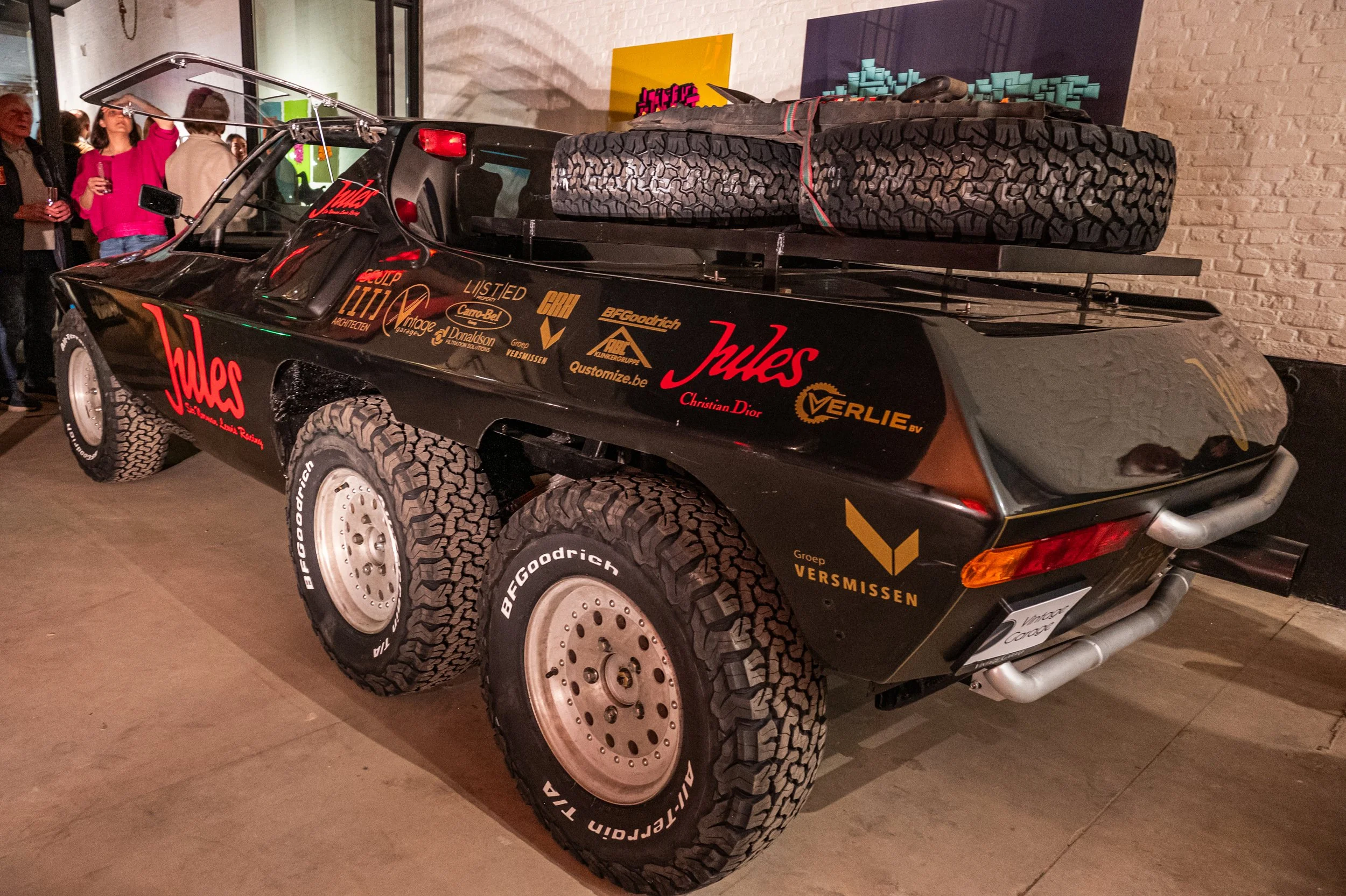 A black rally car with multiple sponsor logos and large off-road tires, on display indoors, with a spare tire and rolled exhaust pipes on the back.