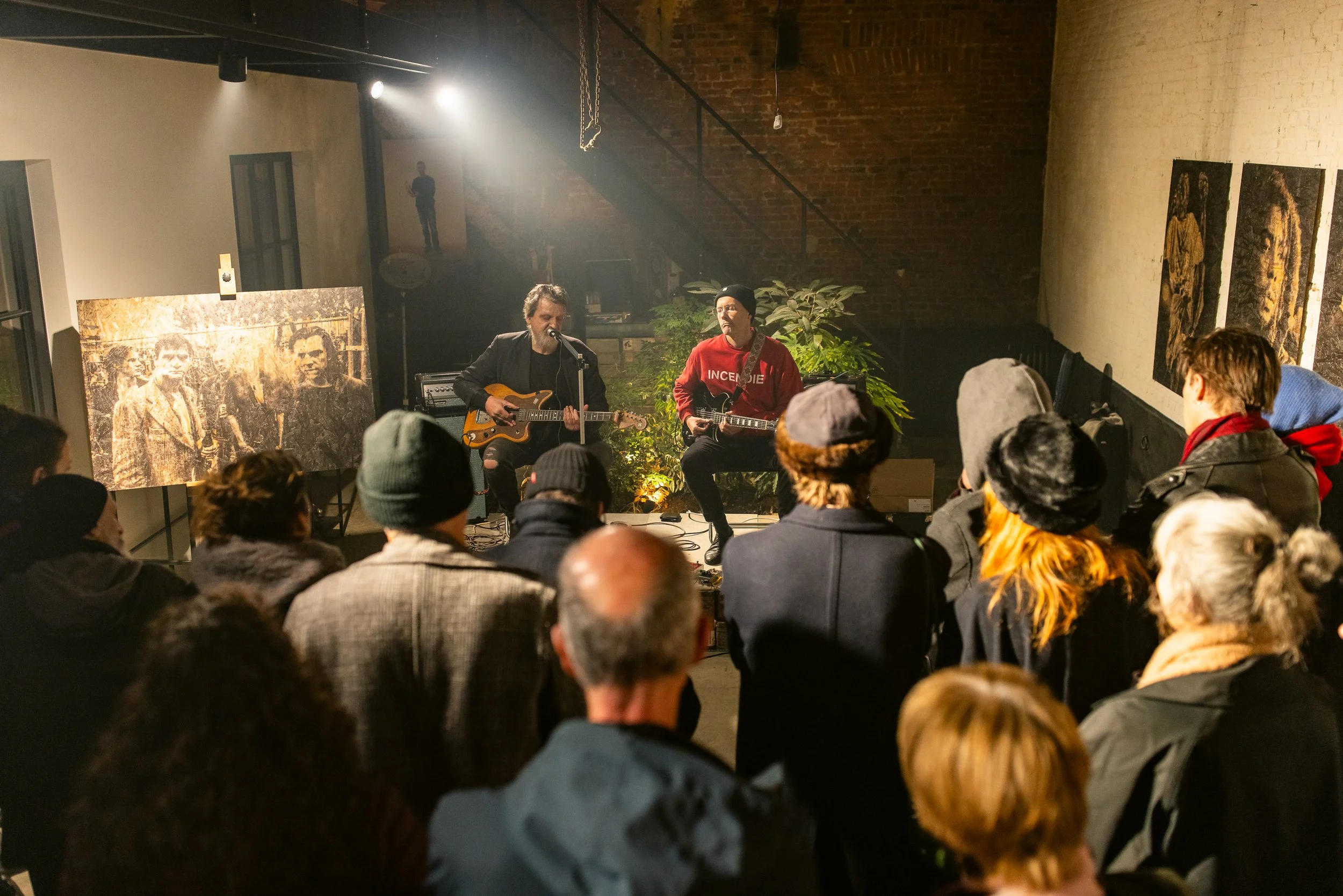 Two musicians perform with guitars on a small stage, surrounded by an audience in an indoor venue with exposed brick and decorated walls.