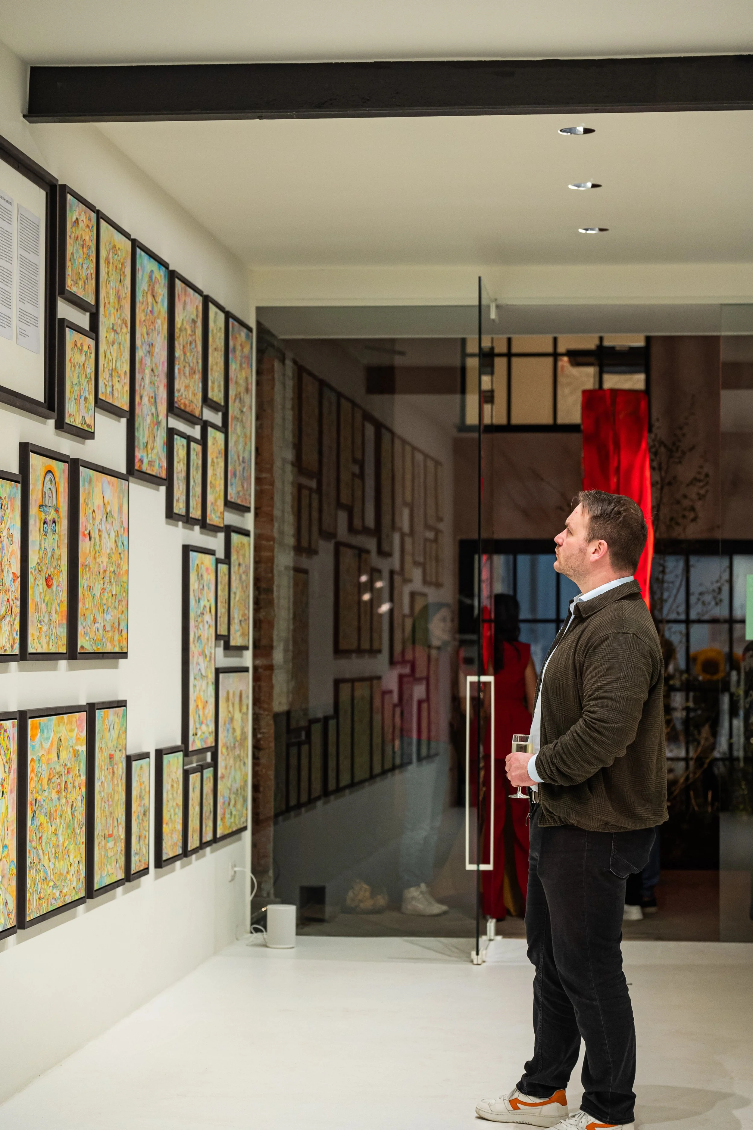 A man dressed in a brown jacket, white shirt, and black pants is standing in an art gallery, observing colorful abstract artwork on the wall while holding a glass of champagne.