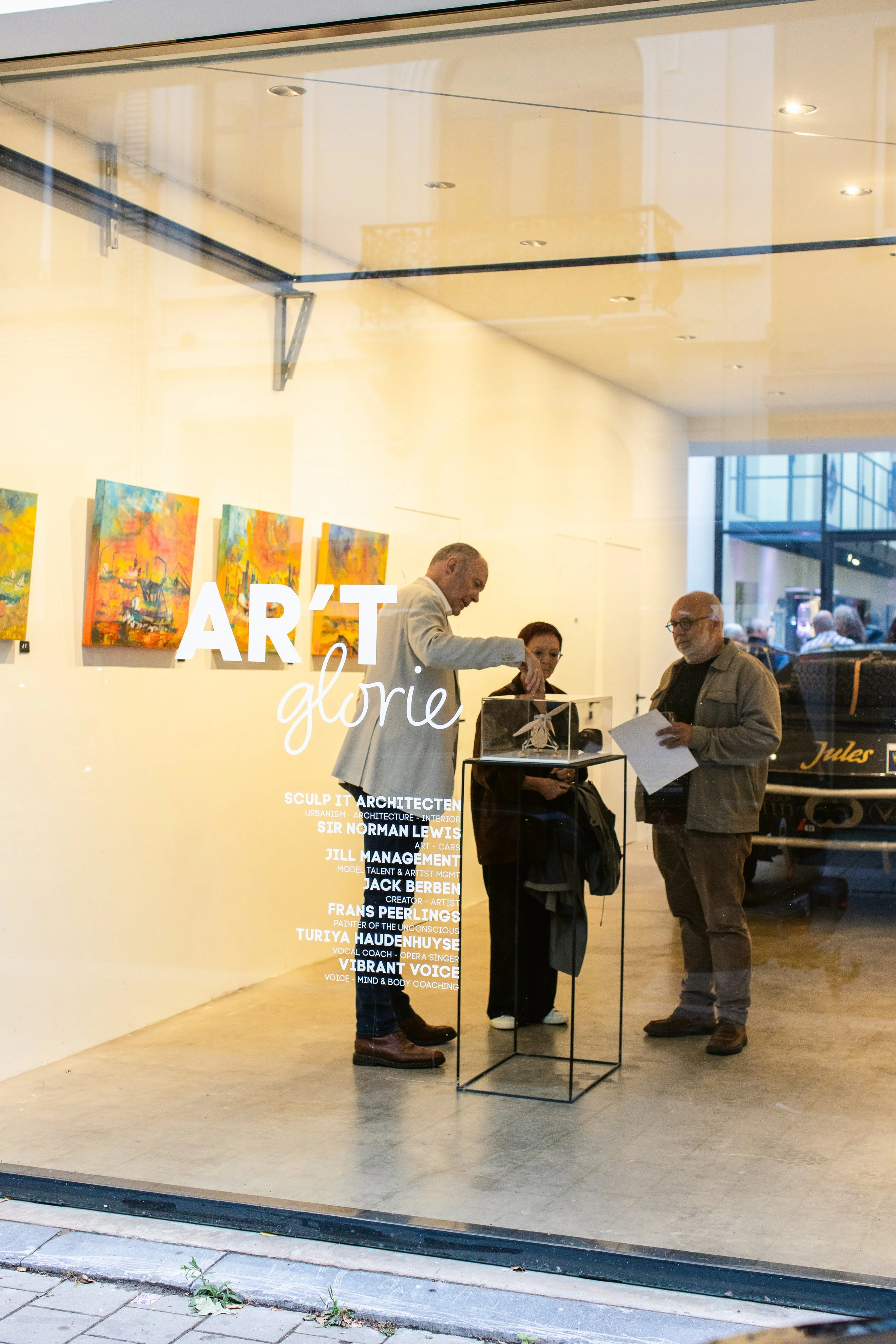 View of an art gallery window with three people inside, examining items on display. The gallery features colorful paintings on the wall and white text on the glass that reads 'ART glorie' with additional smaller text listing artists and their roles.