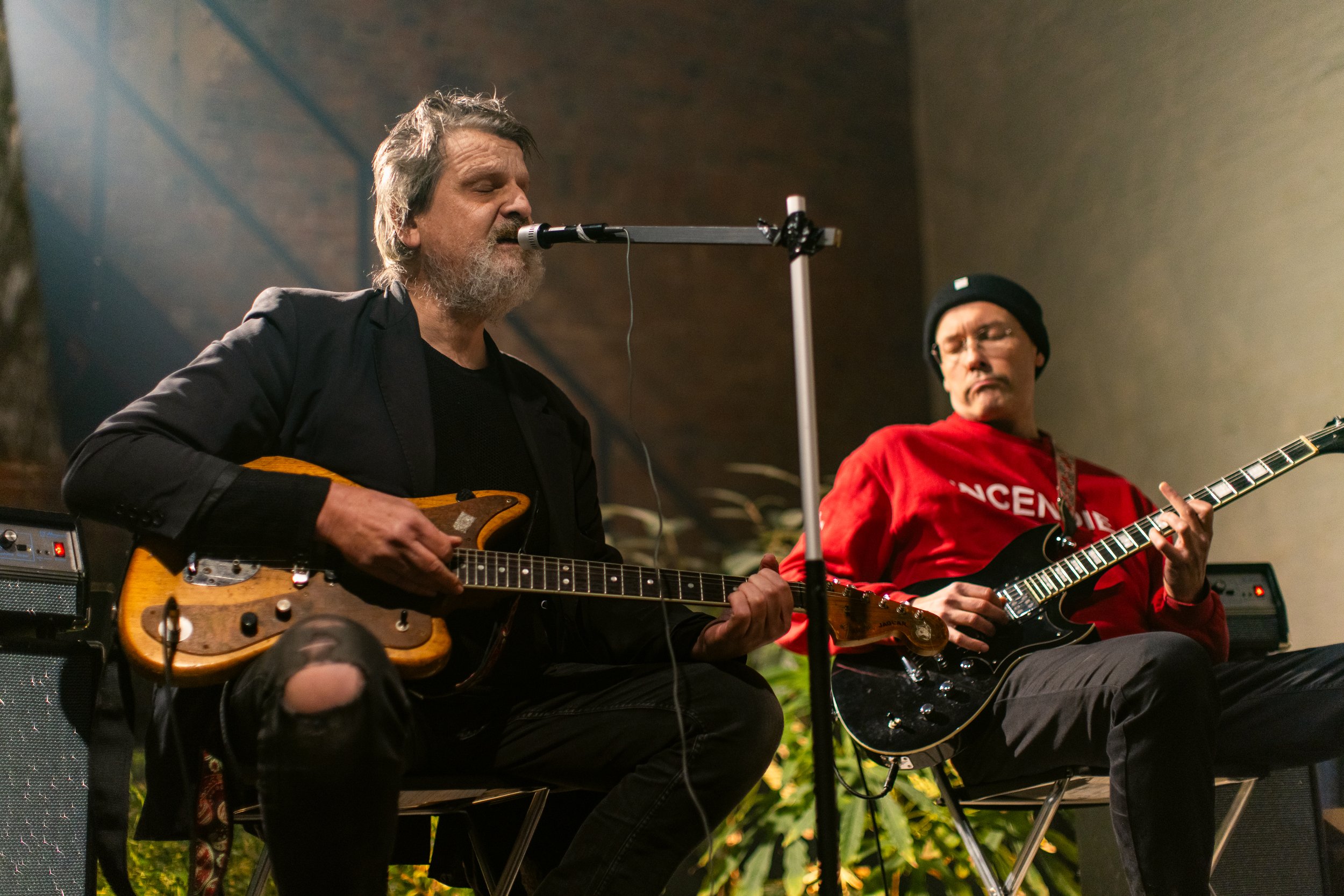 Two men playing guitars and singing into a microphone during a live performance. One man has a beard and is wearing a black jacket, the other man is wearing glasses, a black beanie, and a red hoodie.