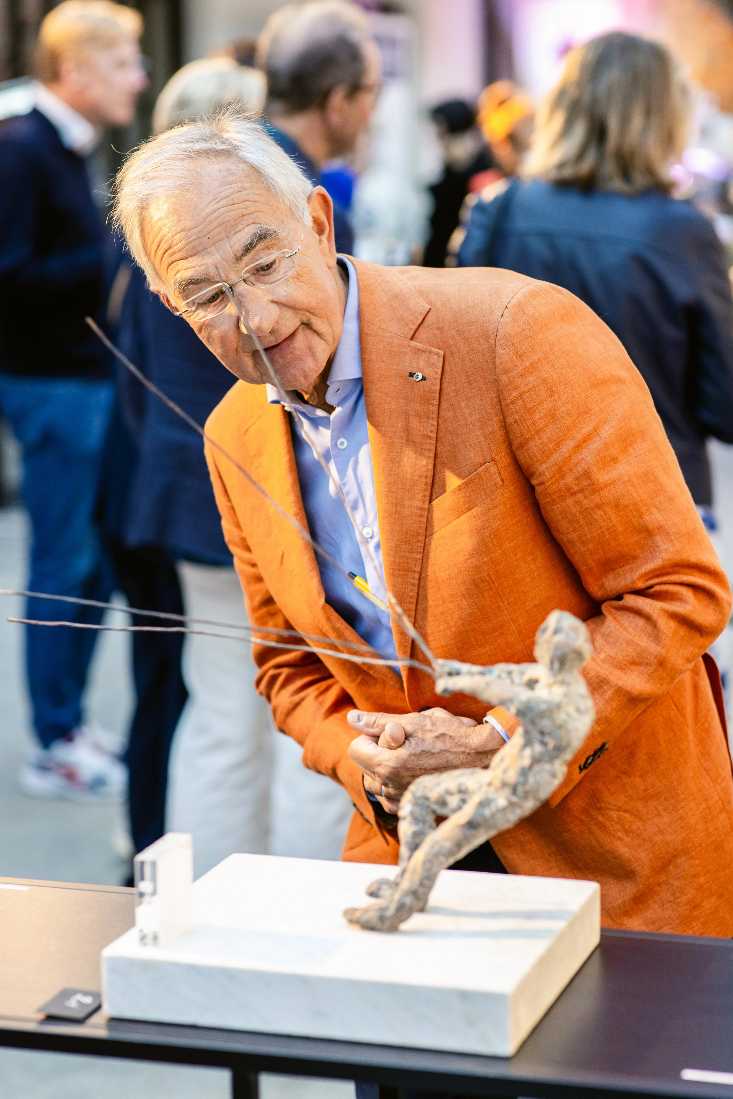 An older man with glasses wearing an orange blazer looking at a sculpture of a person in a crouching position on display. The sculpture is connected by wires, and the man appears intrigued in an indoor gallery setting with other people in the backgro