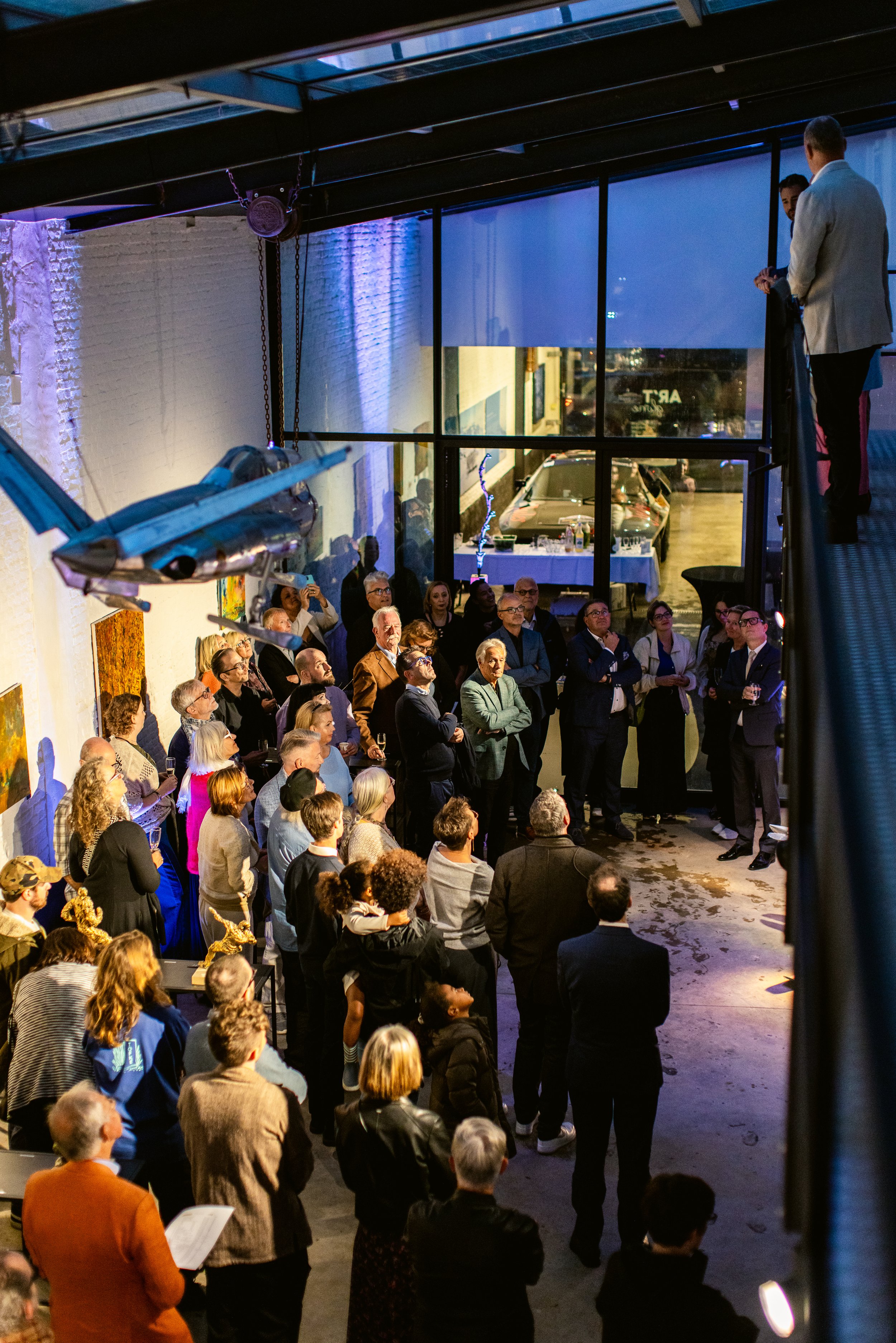A group of people attending an event in a spacious industrial-style venue with artwork on the walls, a model airplane hanging from the ceiling, and a large window showing parked cars outside.