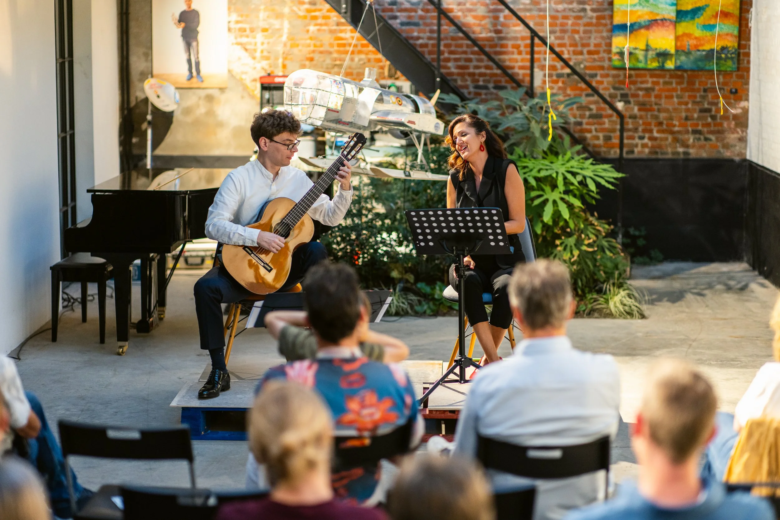 Musicians performing a live guitar and vocals performance in an indoor space with brick walls, greenery, and an audience watching.