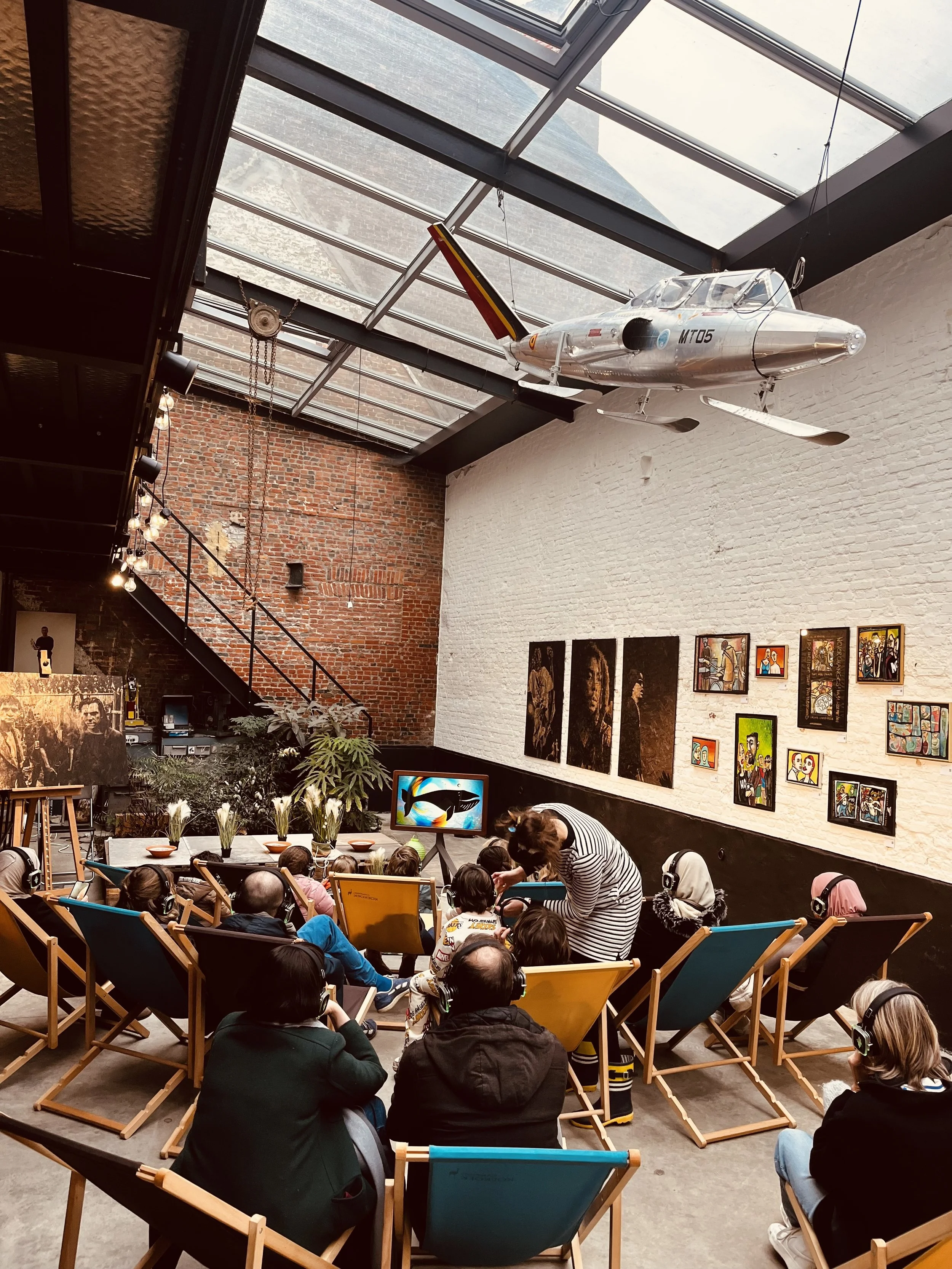 Group of people sitting in an art gallery or museum, wearing headphones and watching a presentation. The gallery has colorful artwork on a white brick wall and a large model airplane hanging from the ceiling.