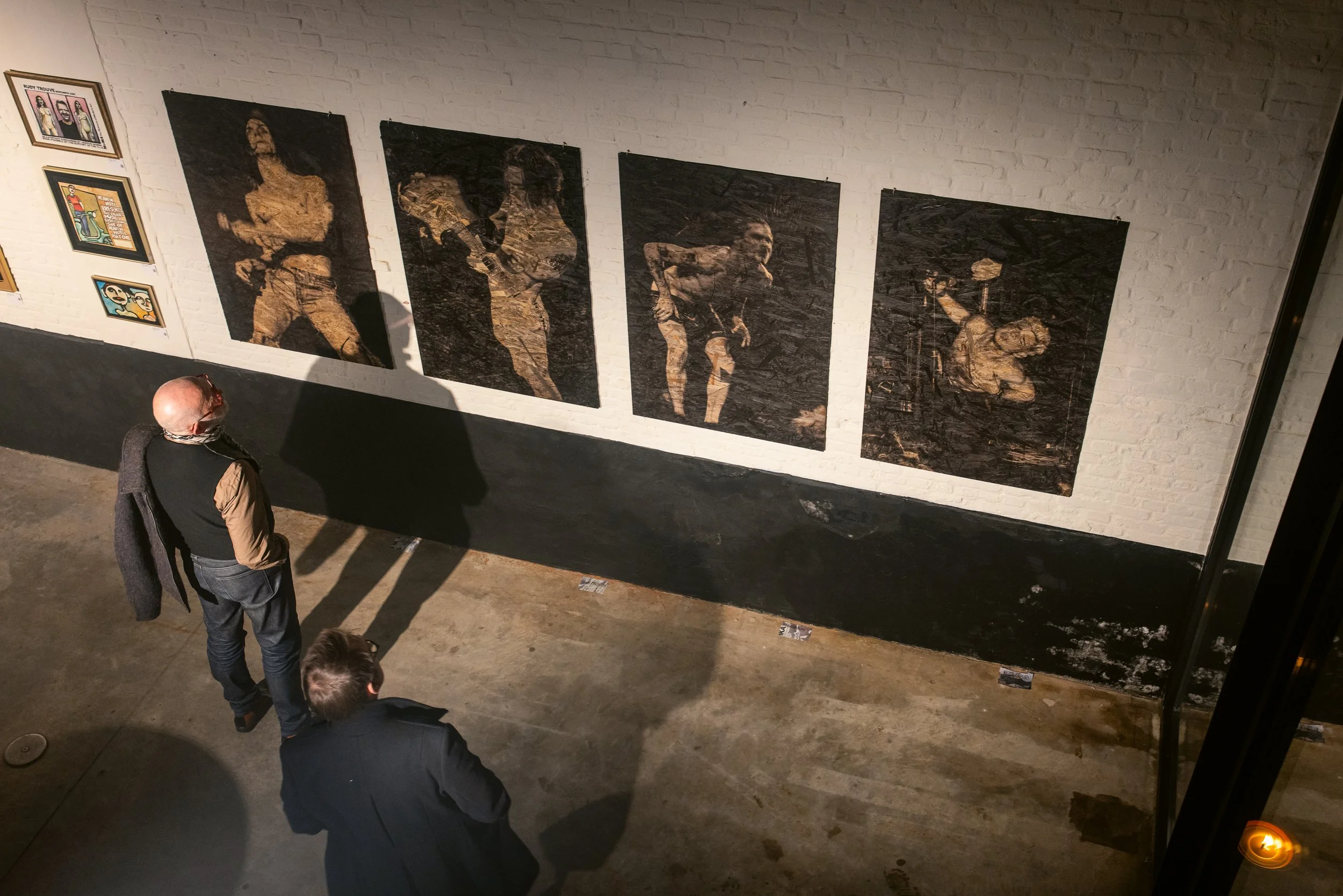 Two people look at a series of four large, dark-toned art pieces on a white brick wall in an art gallery. The artworks depict semi-abstract human figures.