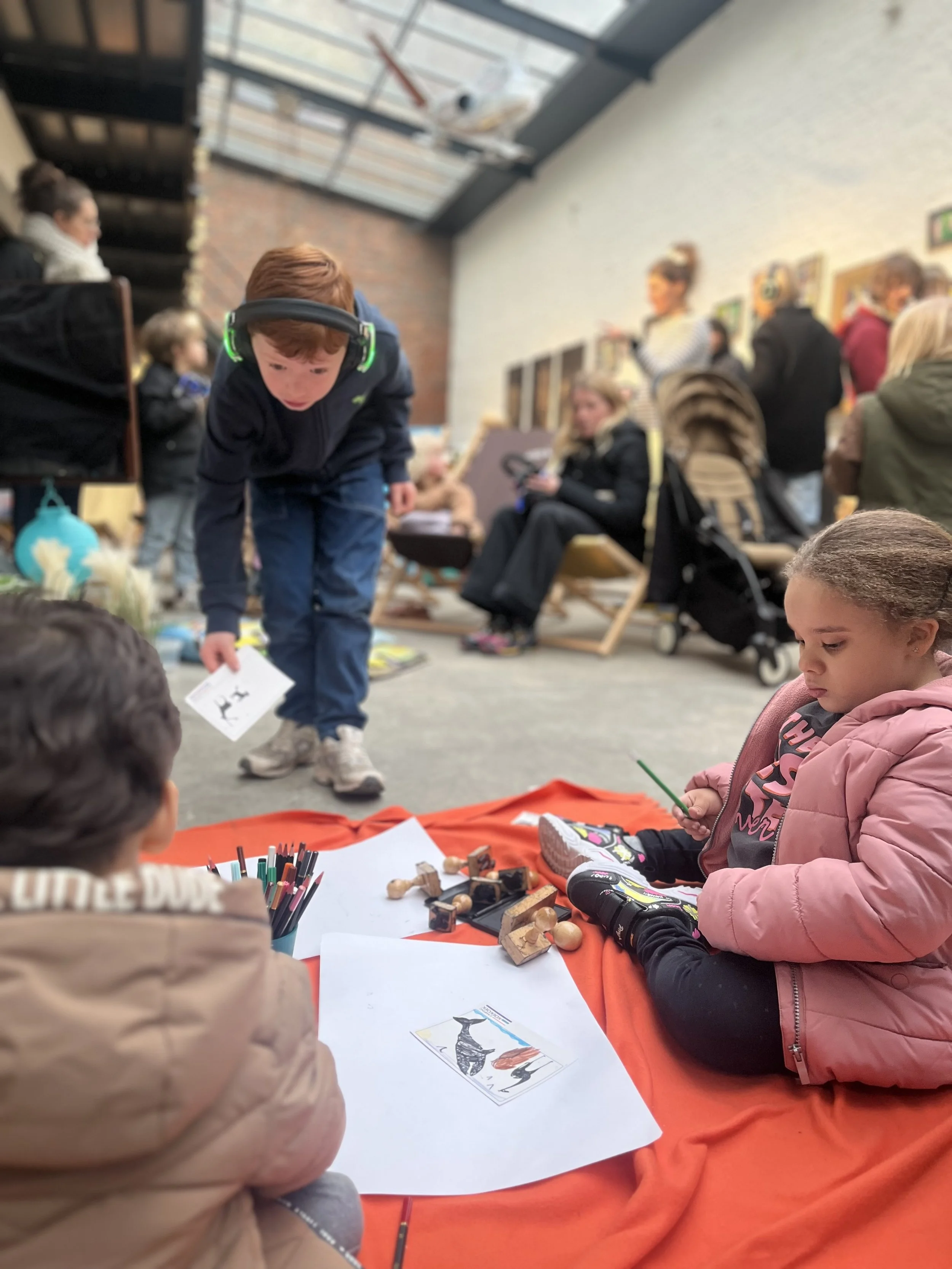 Children sitting on an orange cloth on the floor with toys and coloring pages, while an older boy wearing headphones leans over to look at the items. Adults and other children are in the background, some sitting and some standing, in an indoor space 