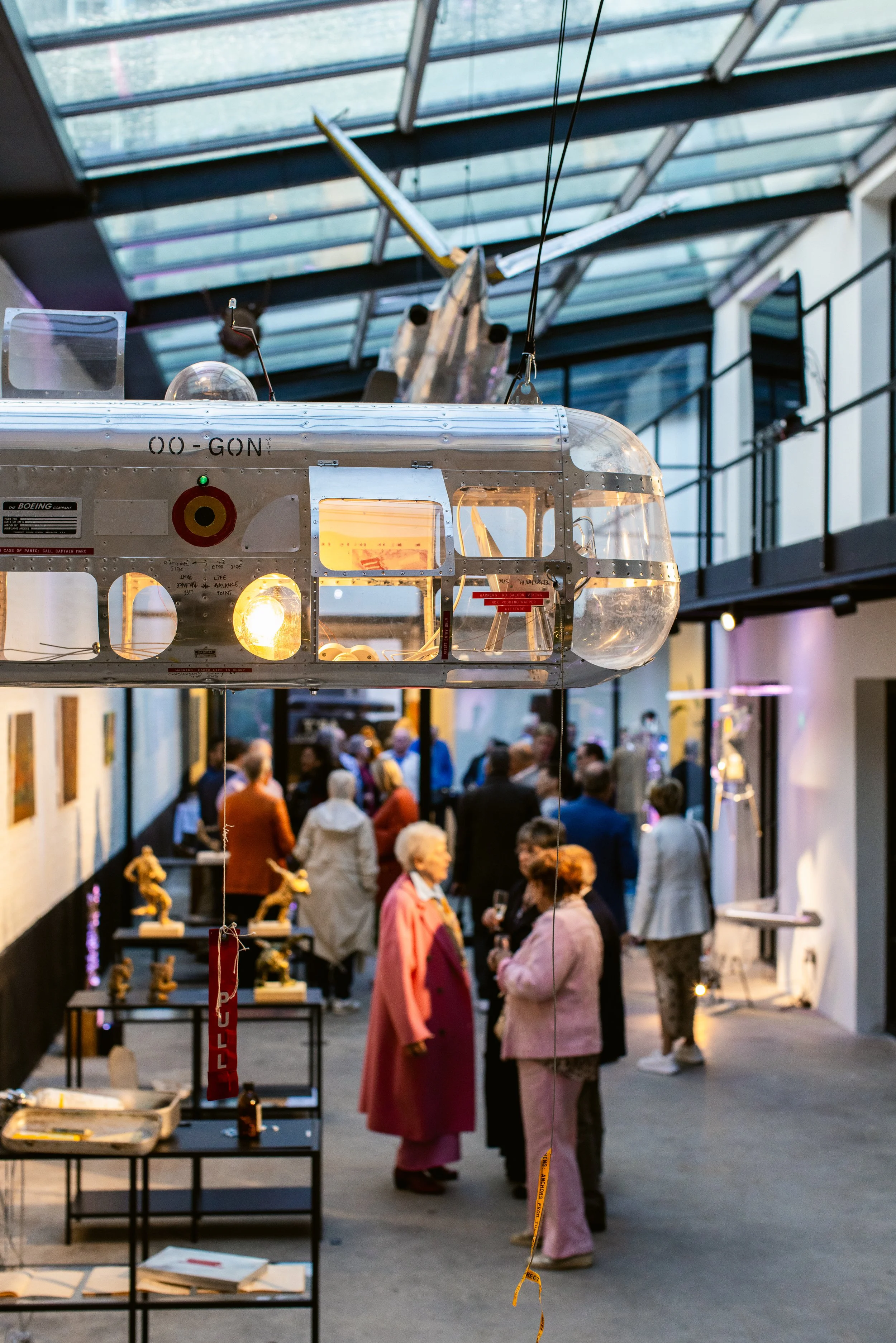 A miniature helicopter hanging from the ceiling inside an art gallery, with people mingling and viewing artwork in the background.