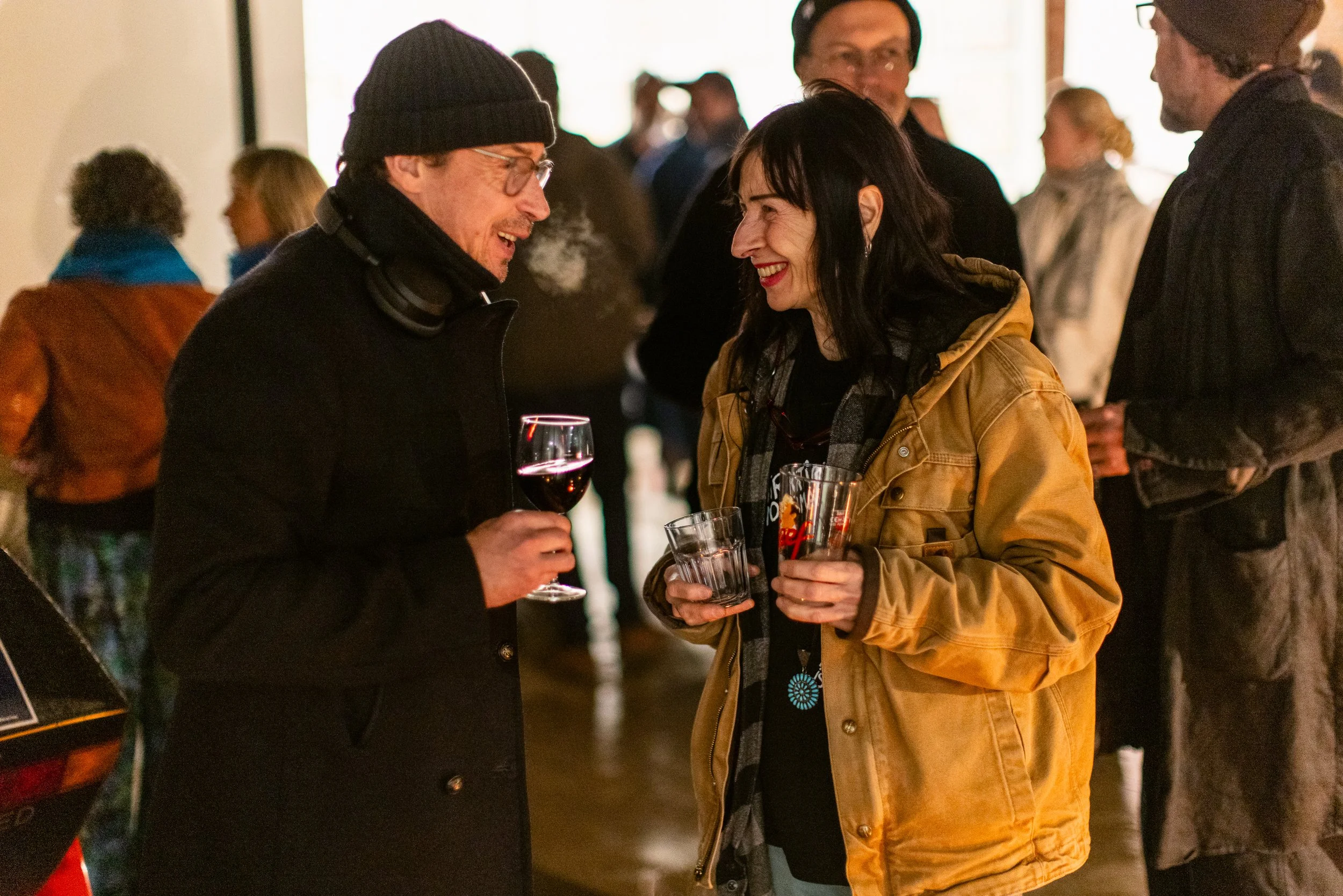 Two people smiling and talking at a social gathering, one holding a glass of red wine, the other holding a beer in a glass. They are in a warmly lit indoor space with several other people in the background.