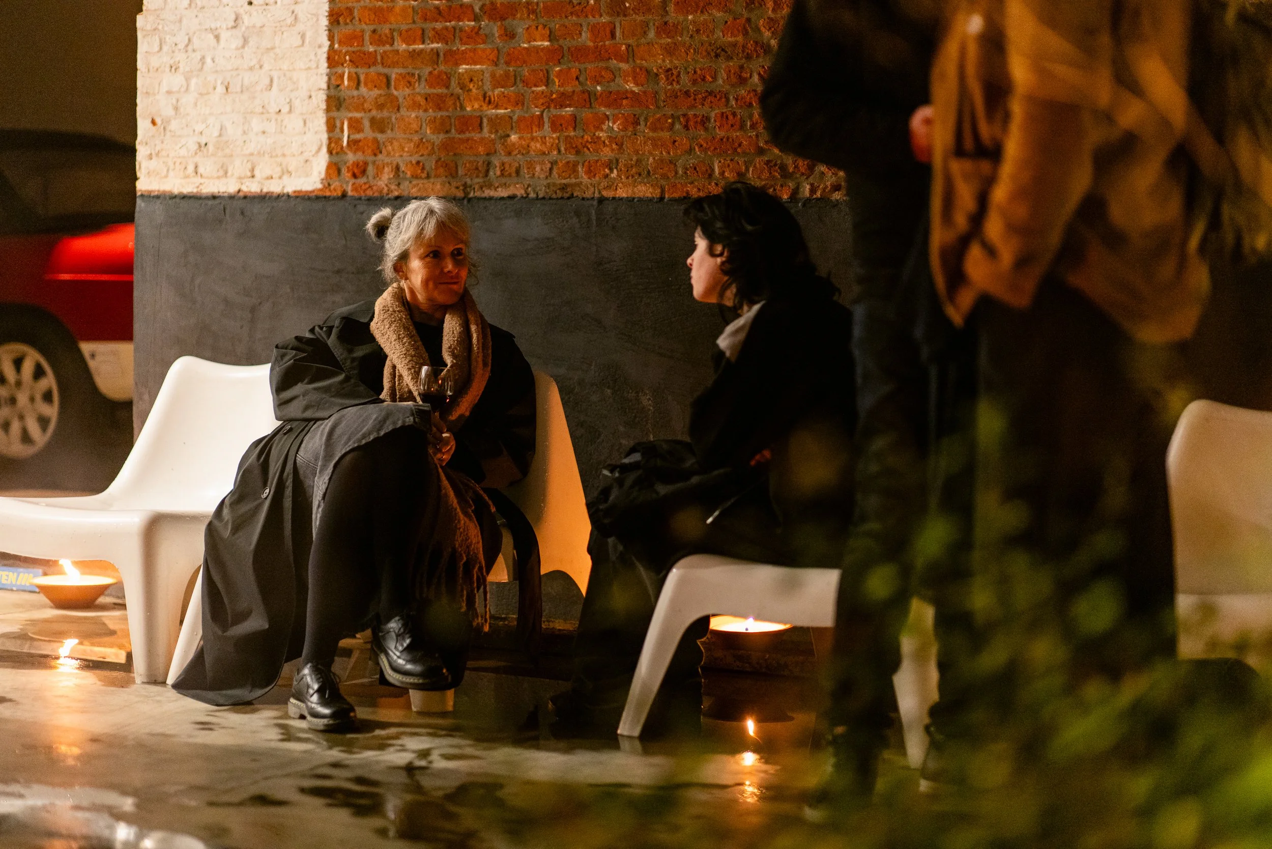 Two women sitting on white benches outside at night, engaged in conversation. One woman is holding a glass of red wine, wearing a black coat and a tan scarf. The other woman has black hair, wearing a dark coat with her arms crossed. A brick wall and 