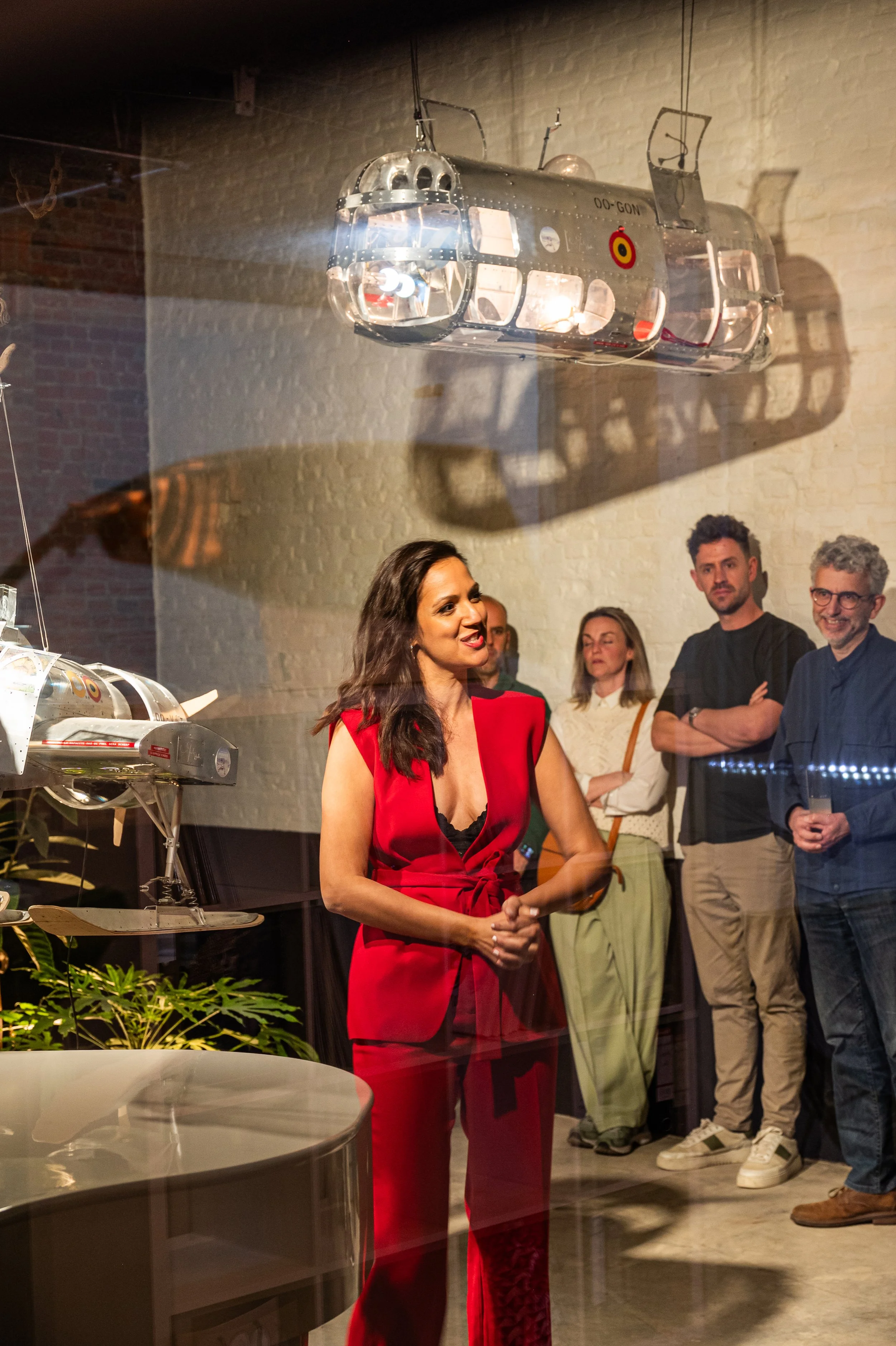 Group of people gathered at an exhibition, viewing scale models of aircrafts displayed behind a glass window, with a woman in a red outfit speaking.