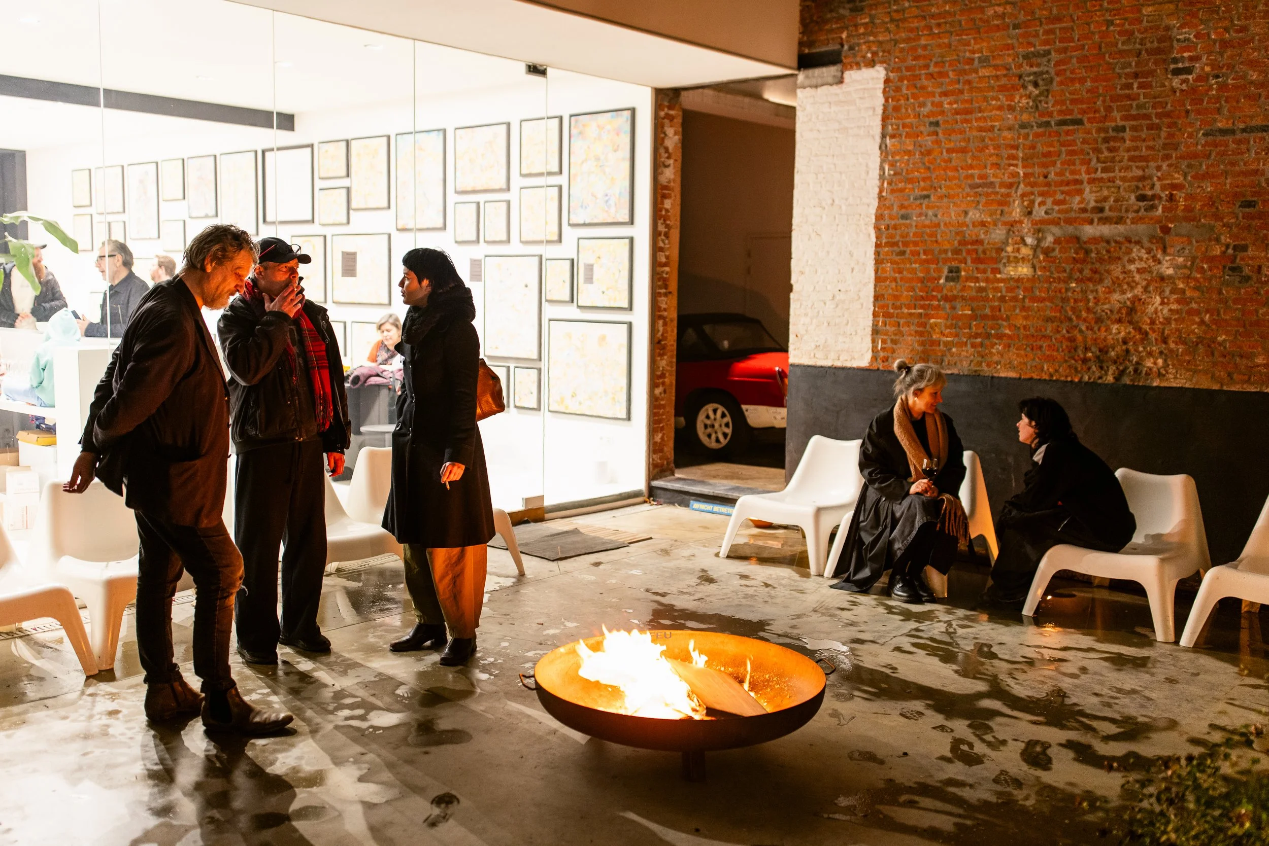 People gathered around a fire pit outside a gallery or art space, with some sitting on chairs and others standing and conversing, during evening hours.