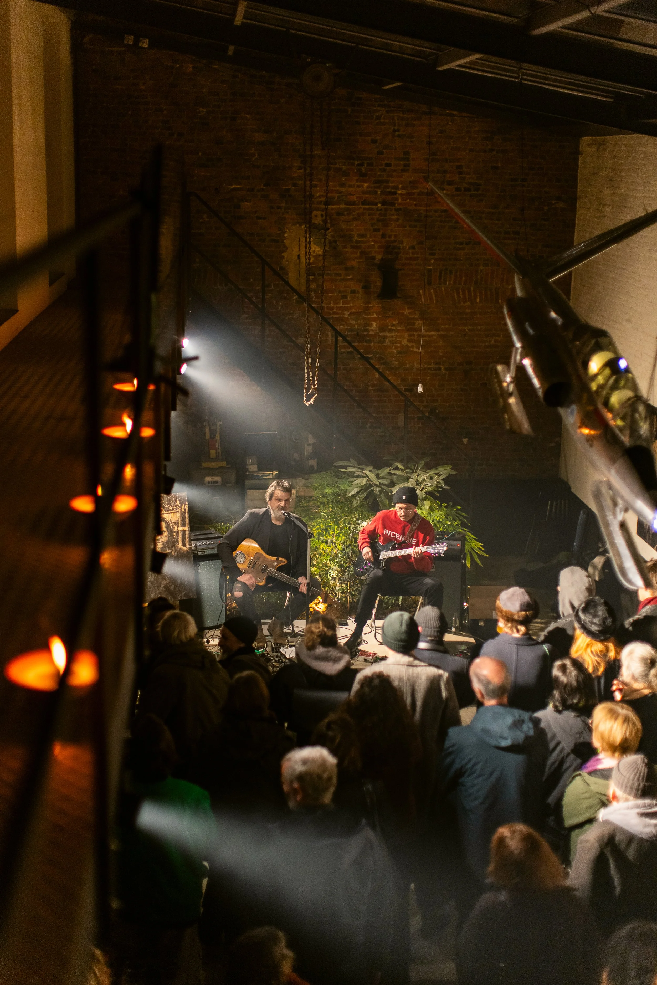 Two musicians performing on stage with a crowd watching, set in a venue with brick walls and plants as decoration.