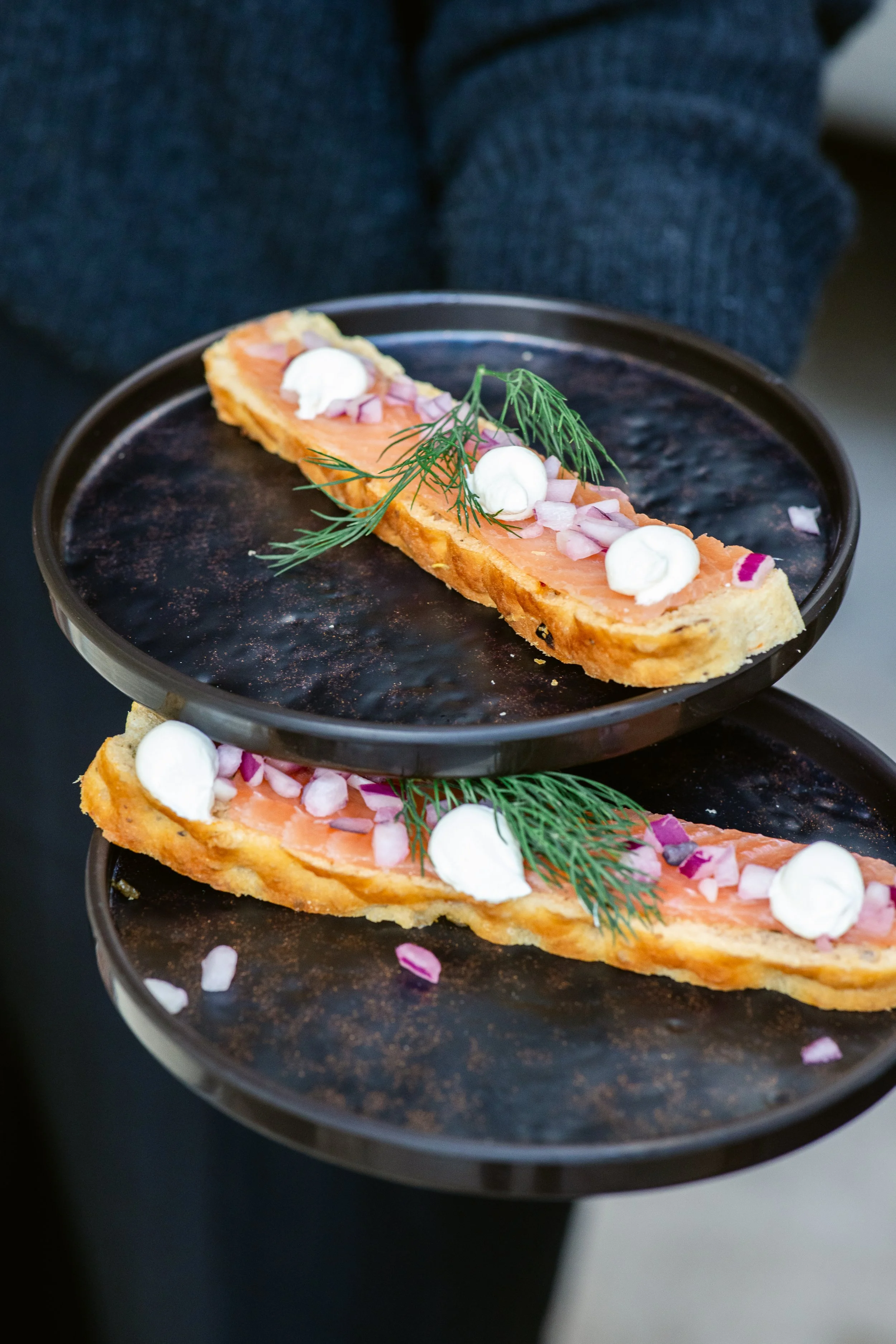 Two trays of smoked salmon on bread, garnished with dill, chopped onions, dollops of sour cream, and pink salt flakes.