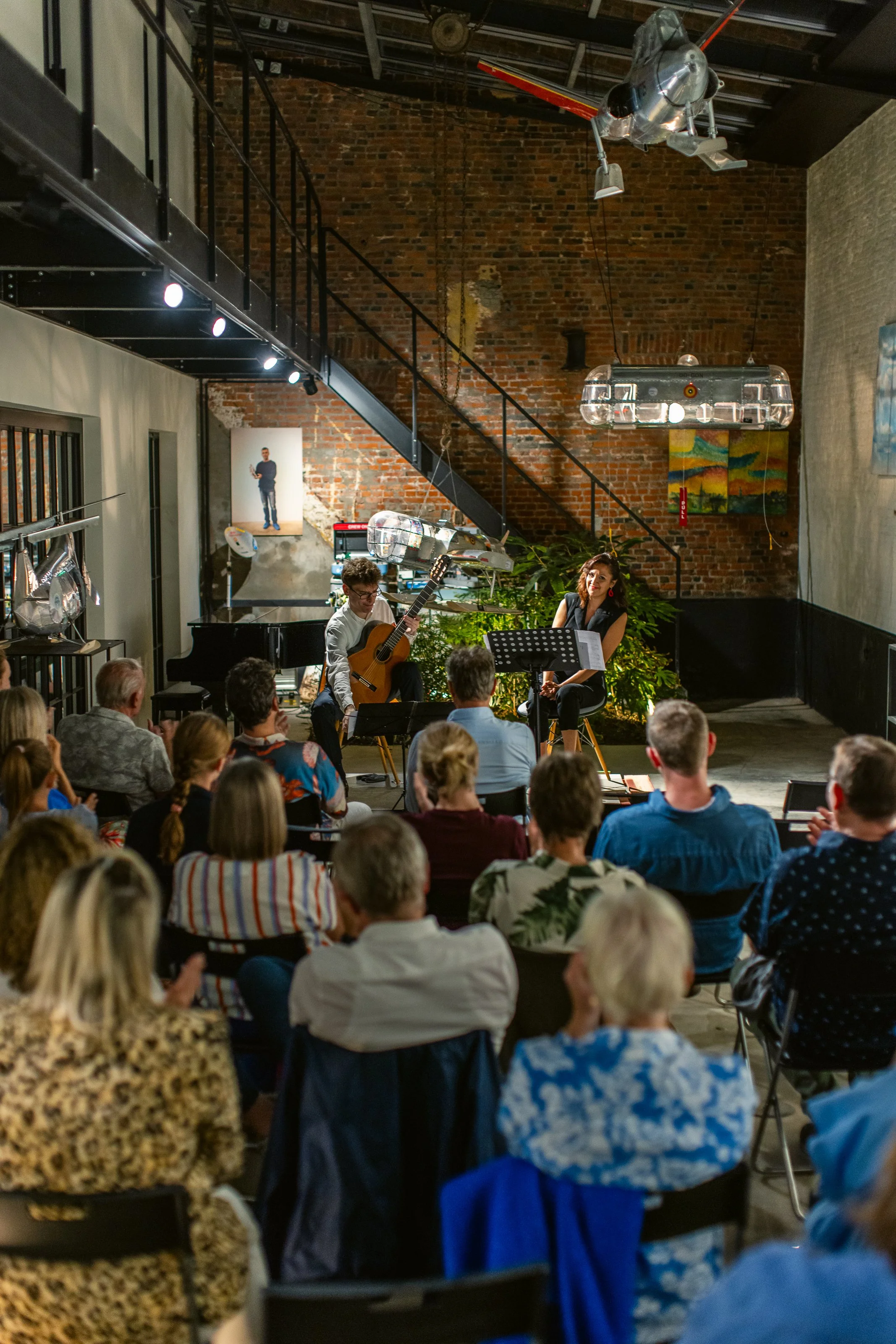 Musicians performing on stage with audience watching in a cozy, indoor venue with brick walls and hanging modern lights.