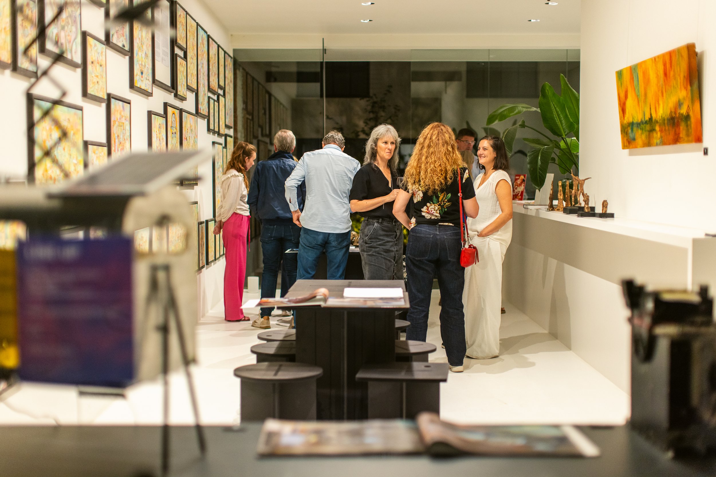 People viewing artwork in a modern art gallery, with framed paintings on white walls and sculptures on a white counter.