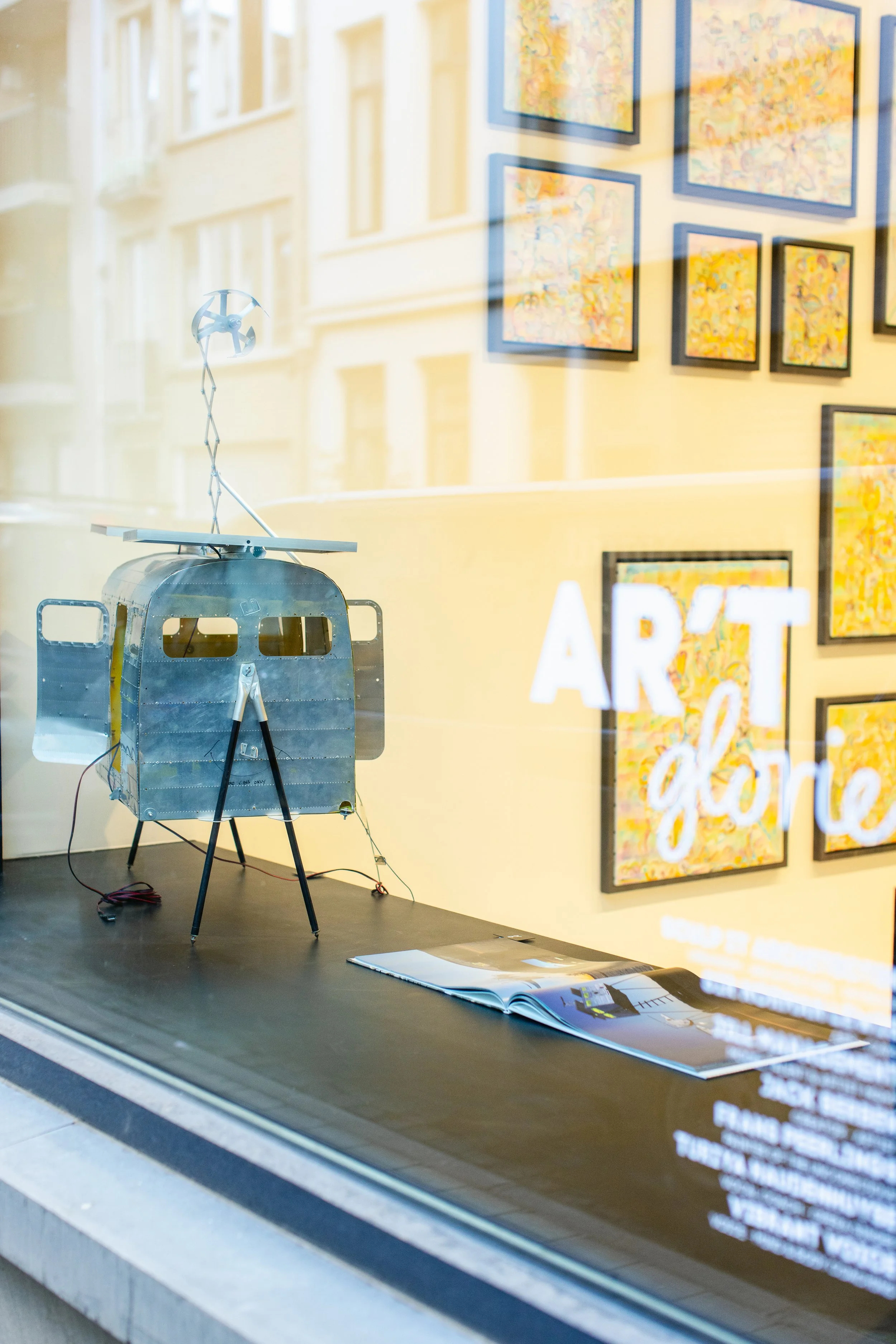 A metallic model of a spacecraft displayed in an art gallery window, with colorful abstract paintings on the yellow wall inside, and a reflection of buildings outside on the glass.
