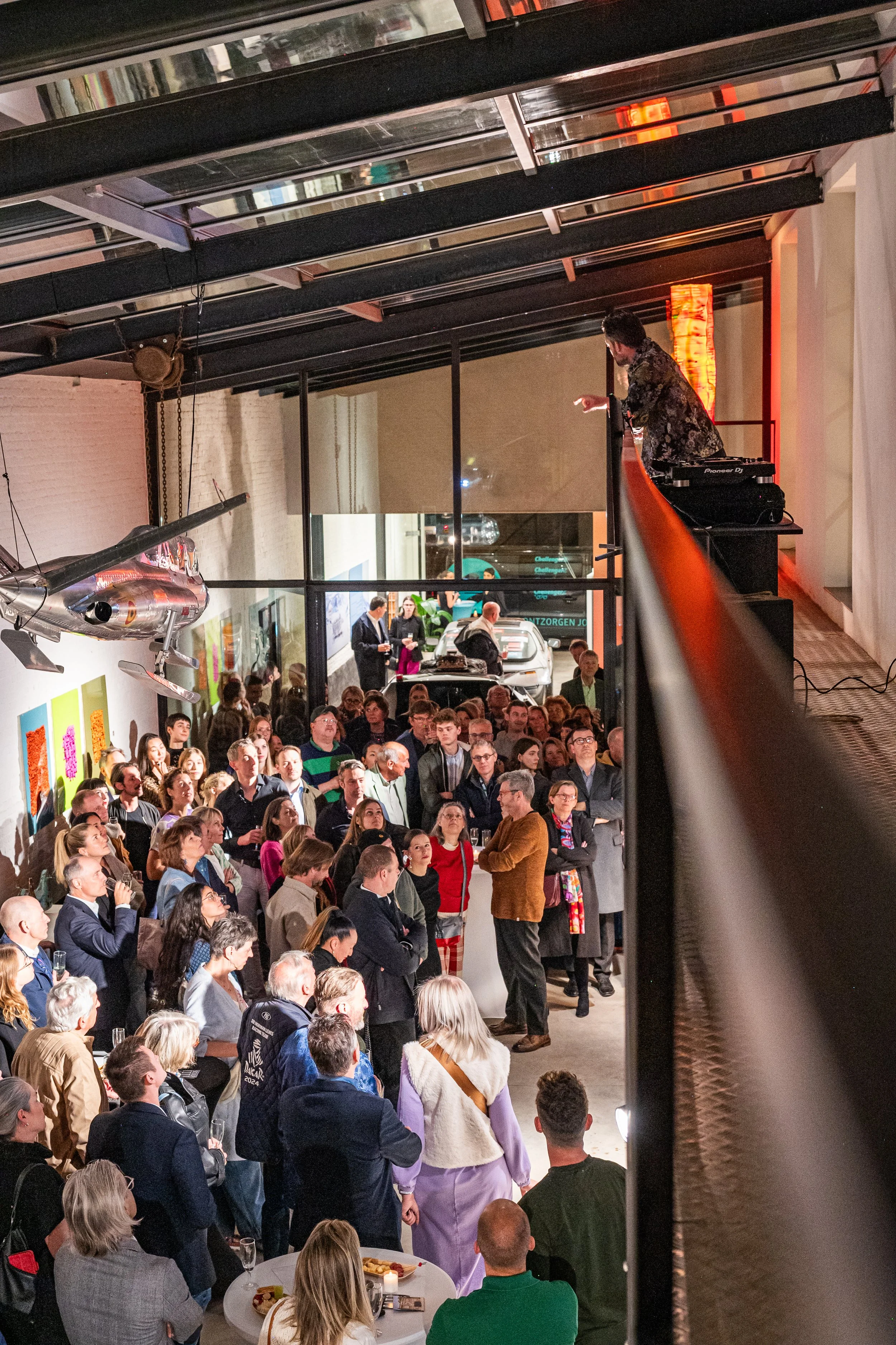 A crowded indoor event space with many people gathered around a speaker. The room has artwork on the wall and model airplanes hanging from the ceiling, with one person standing near a railing above observing the crowd.