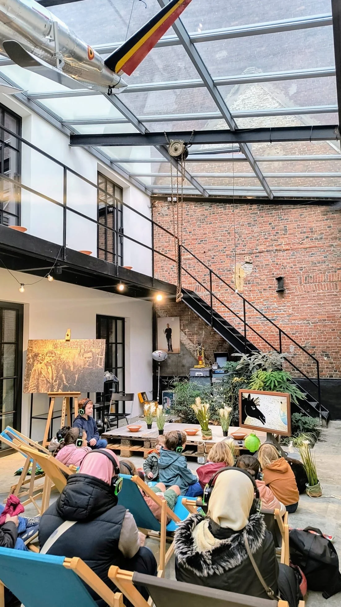Children sitting on deck chairs and floor, listening to a storyteller in an art gallery or museum, with paintings and plants, an exposed brick wall, and a glass ceiling with a model airplane hanging.