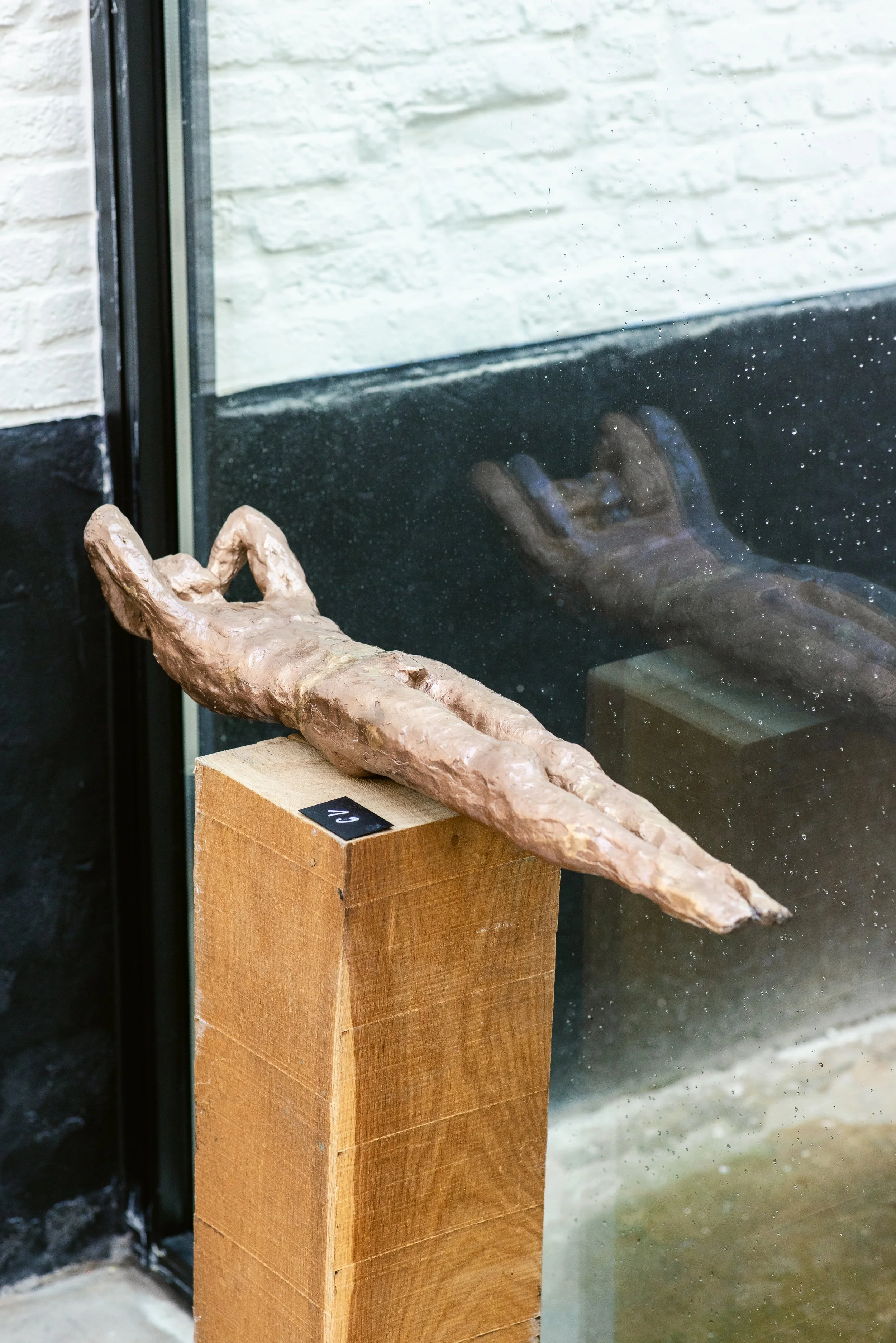 A bronze sculpture of a reclining human figure with one arm raised, displayed on a wooden pedestal in front of a glass window.