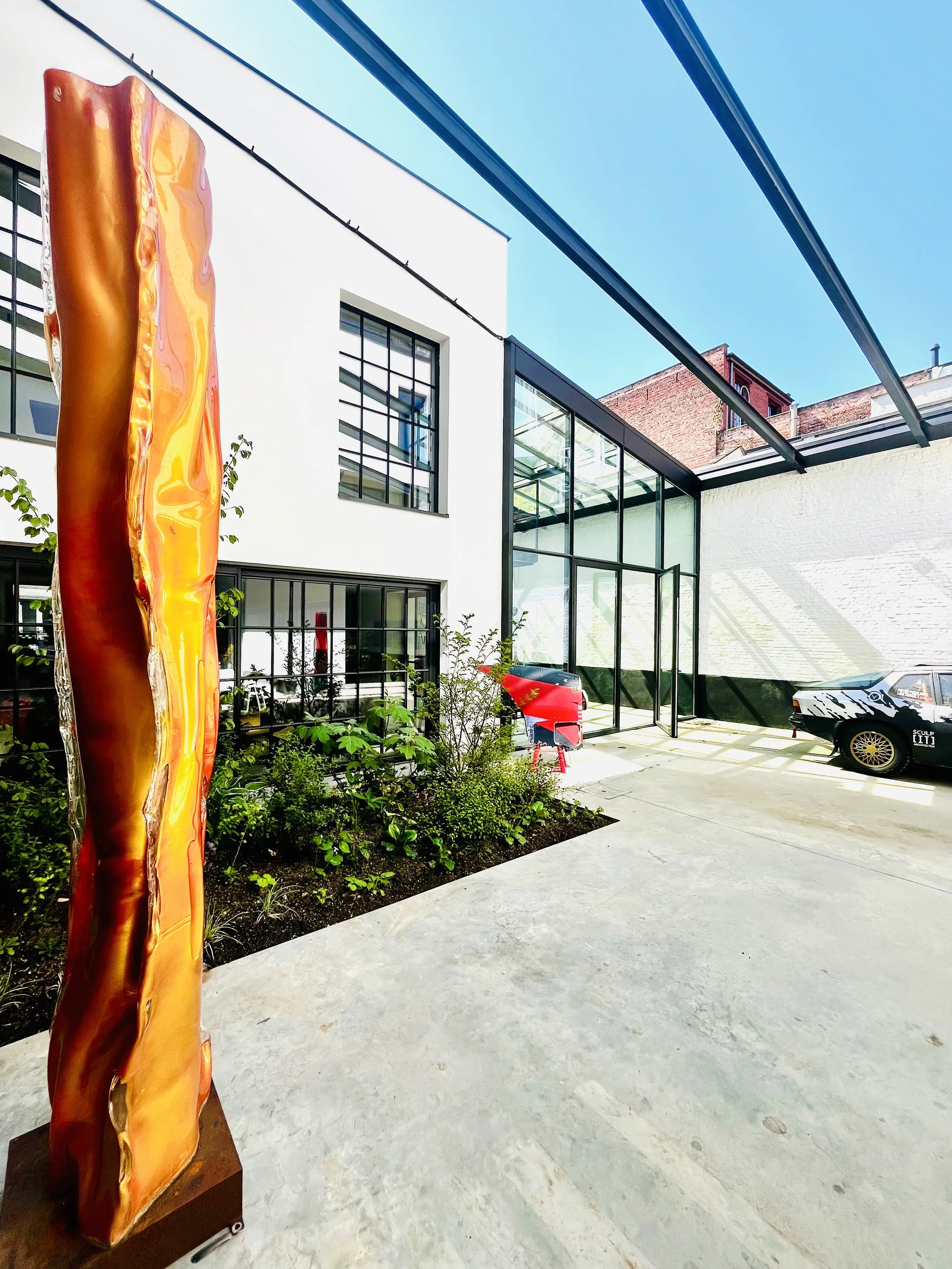 Contemporary art sculpture resembling a molten metal slab outdoors, adjacent to a modern building with glass and white walls, parking lot with cars, and a clear blue sky.