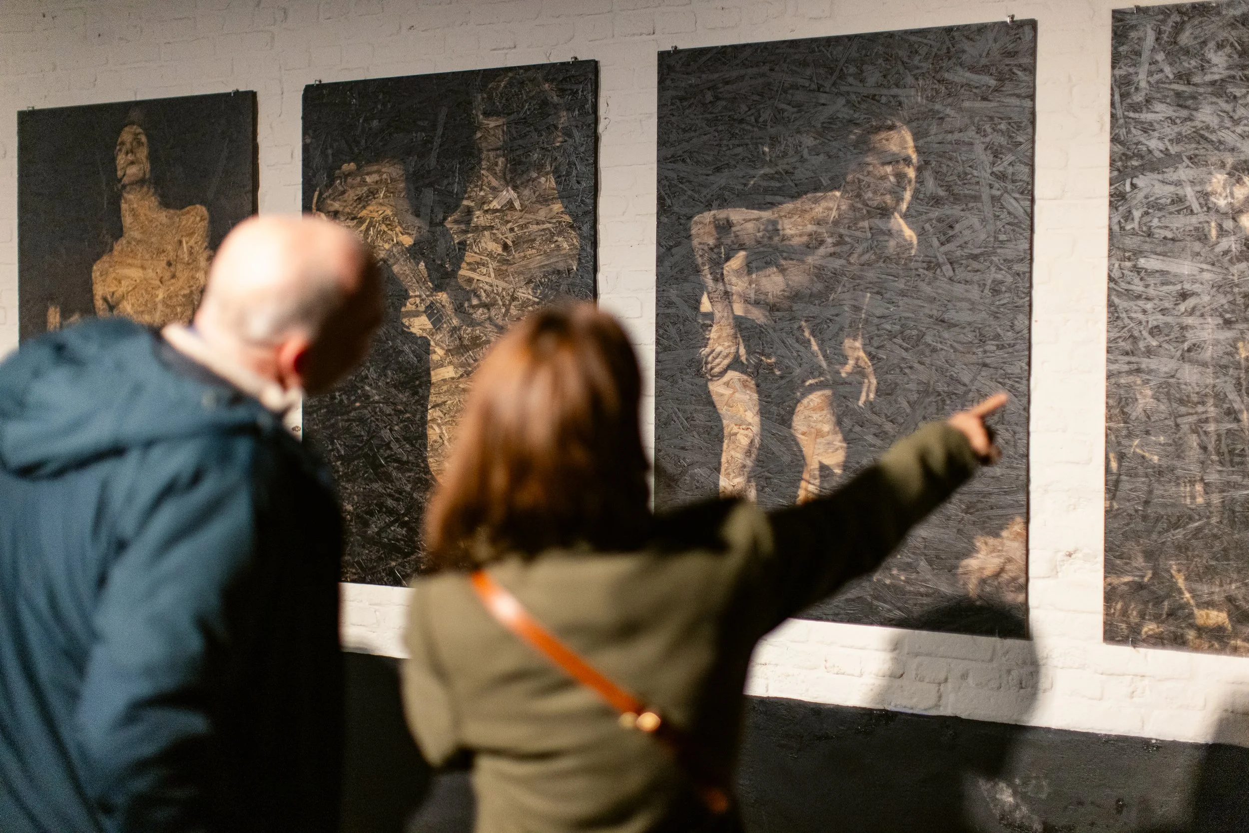 Two people, one man and one woman, viewing artwork on a gallery wall. The artwork appears to be mixed media or textured paintings of women, displayed on a white brick wall.