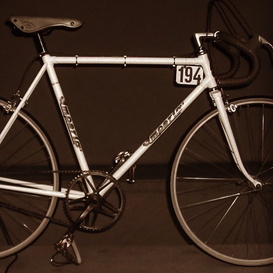 Bicicletta da corsa d'epoca bianca esposta contro una parete scura, con numerino 194 appeso al telaio.