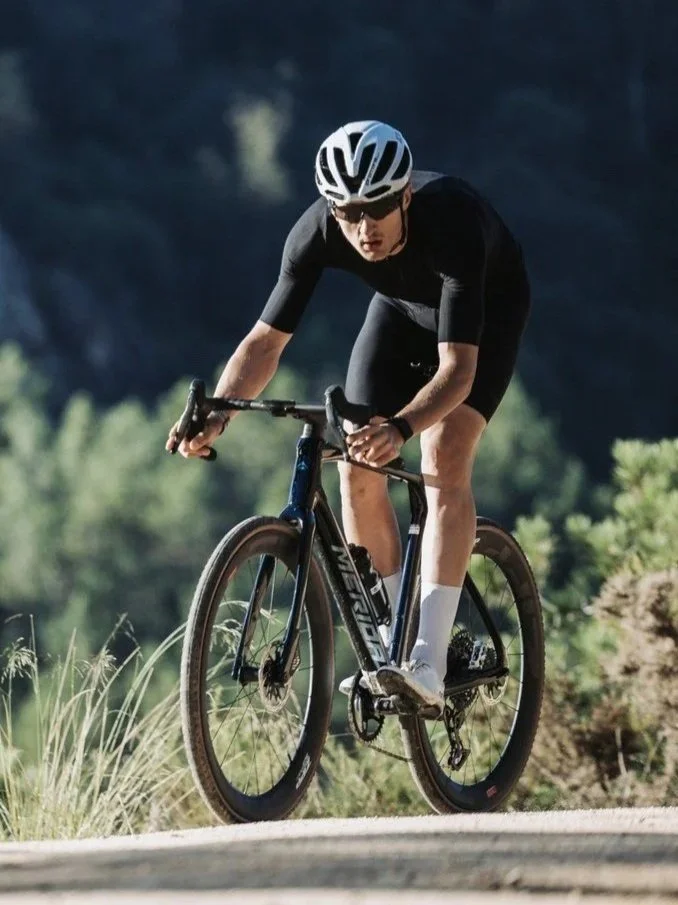 Un ciclista vestito di nero con casco bianco pedala in un paesaggio naturale con vegetazione verde.