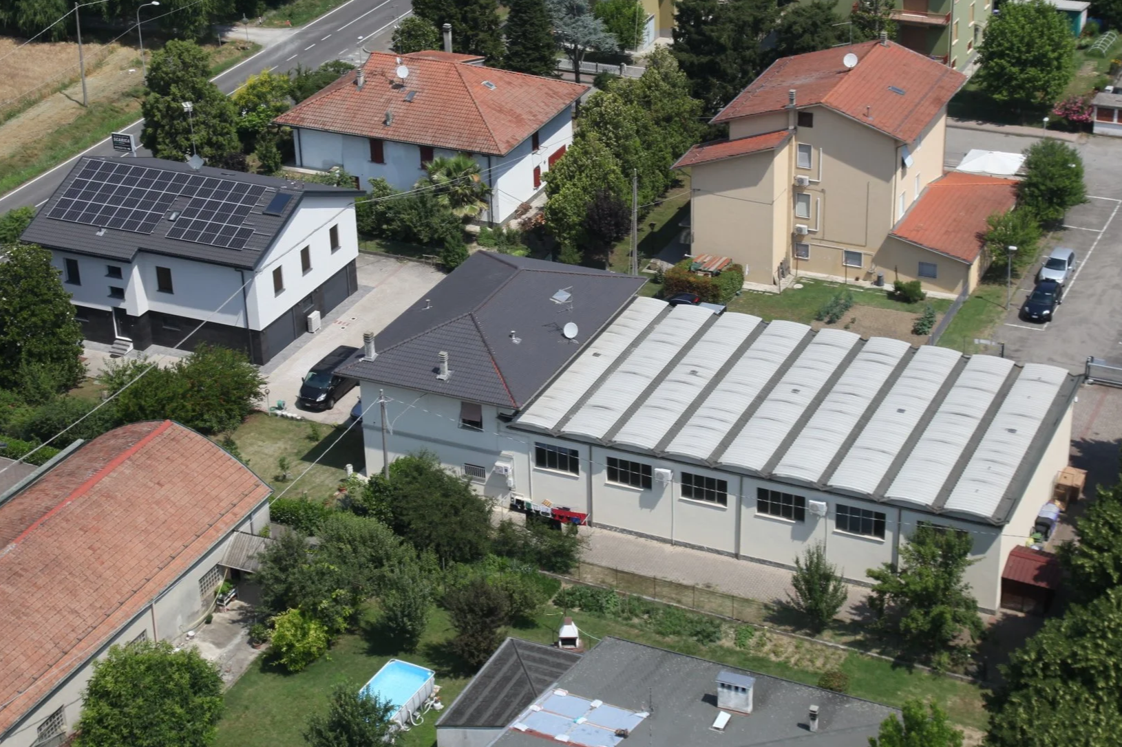 Vista aerea di case residenziali con tetti di diversi stili, tra cui uno con pannelli solari, circondate da alberi e giardini.