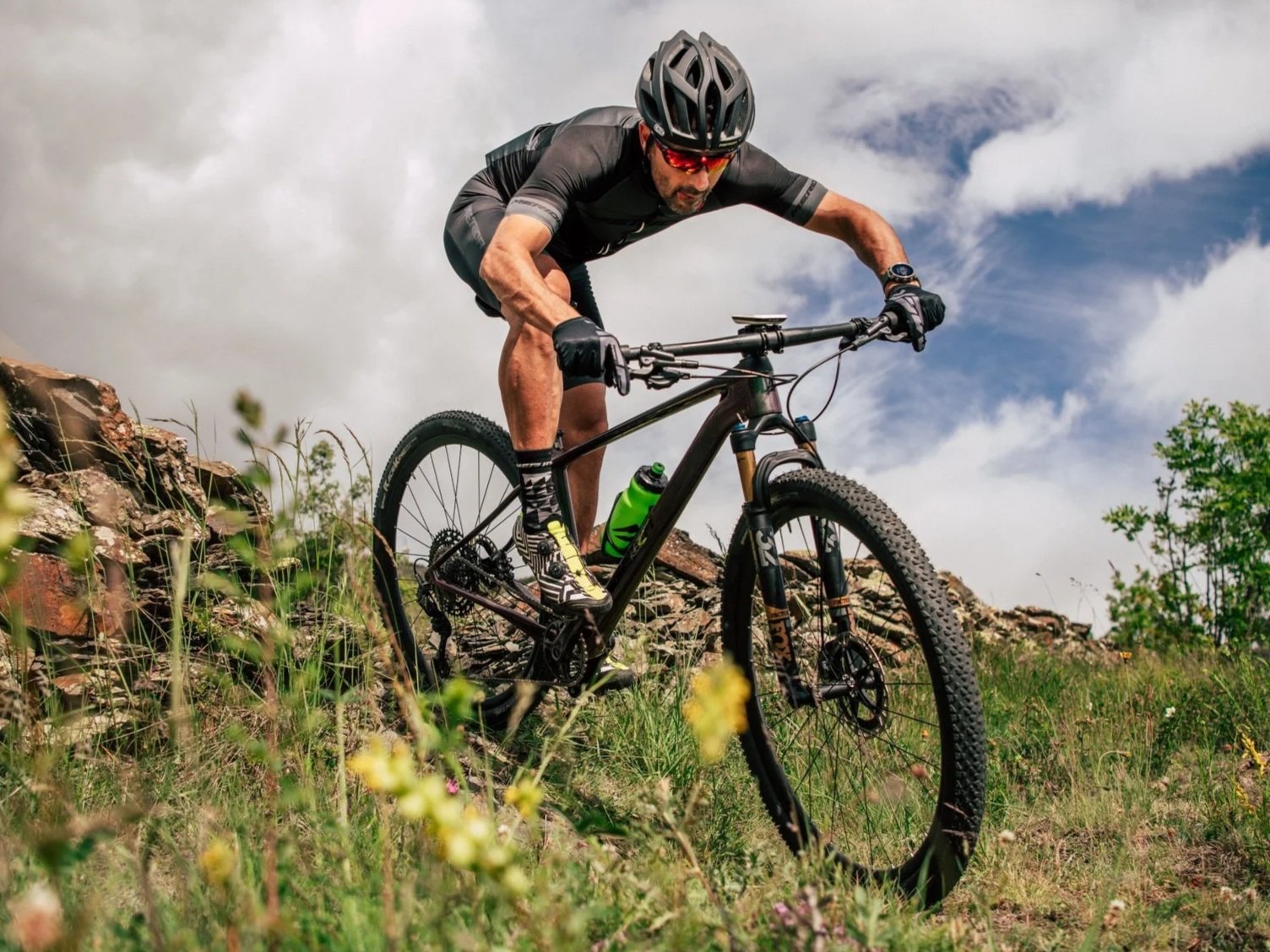 Uomo in bicicletta da montagna su un sentiero con erba e rocce, indossa casco e abbigliamento sportivo, sfondo con nuvole e cielo blu.