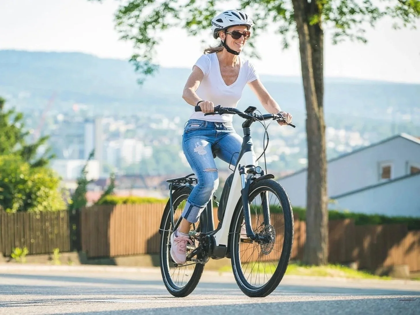 Una donna che pedala su una bicicletta elettrica lungo una strada, indossa casco e occhiali da sole, con un paesaggio urbano e natura sullo sfondo.