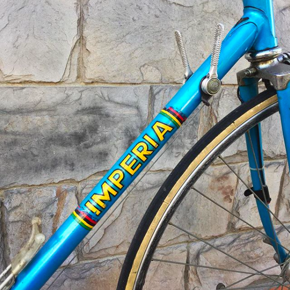 Bicicletta blu con scritta 'IMPERIA' sul telaio, parcheggiata contro un muro di pietra.