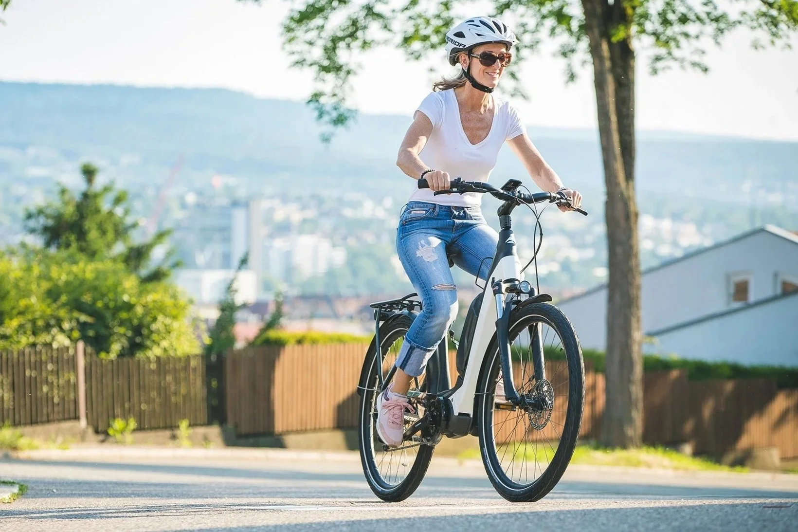 Una donna che pedala in bicicletta su una strada all'aperto in una giornata di sole, indossando casco e occhiali da sole, con sfondo di alberi e case.