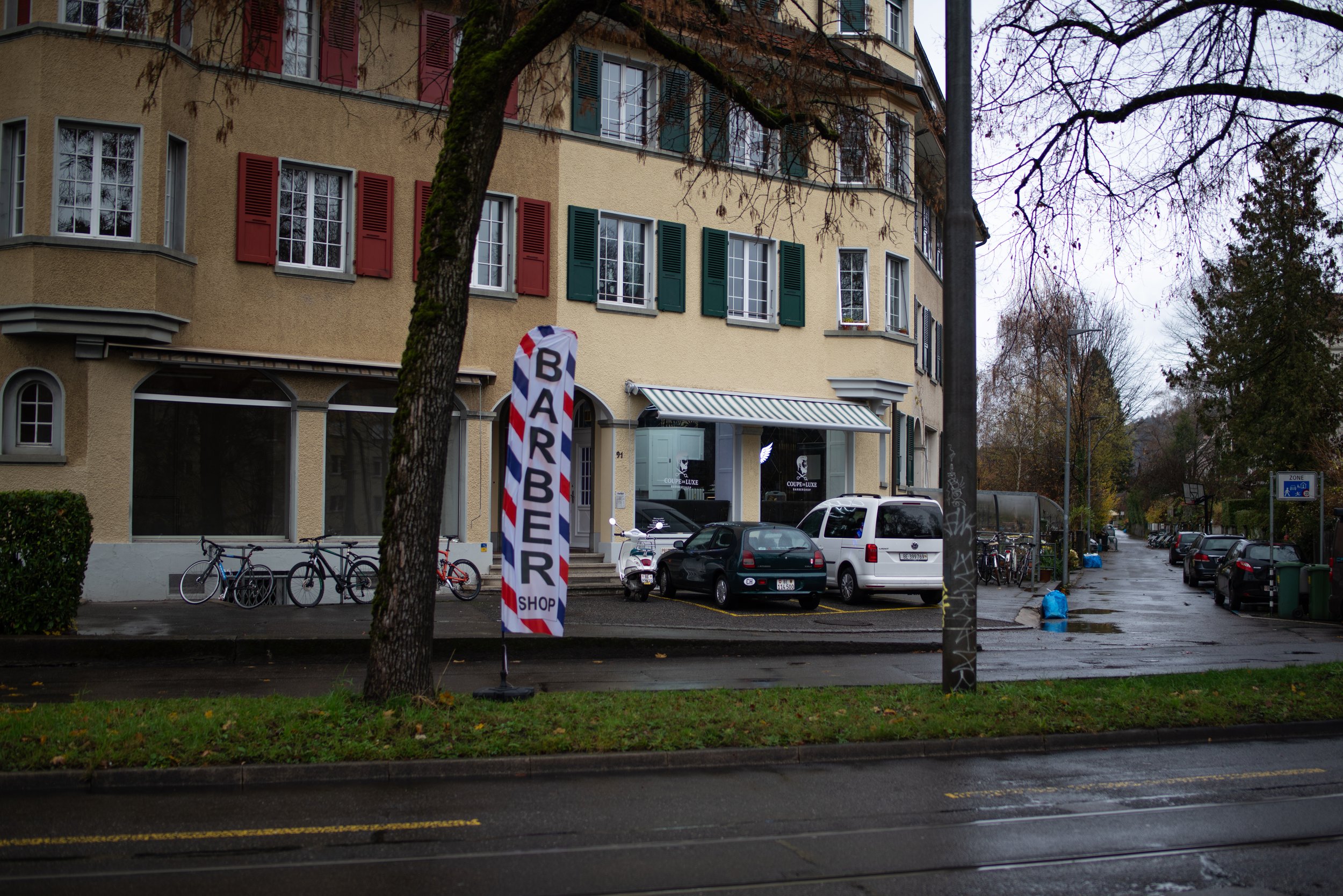 Straßenszene mit Parkplätzen, Bäumen, mehrstöckigem Gebäude mit grünen und roten Fensterläden, Fahrradständern und einem Schild mit der Aufschrift "BARBER SHOP" auf einem Fahnenmast.