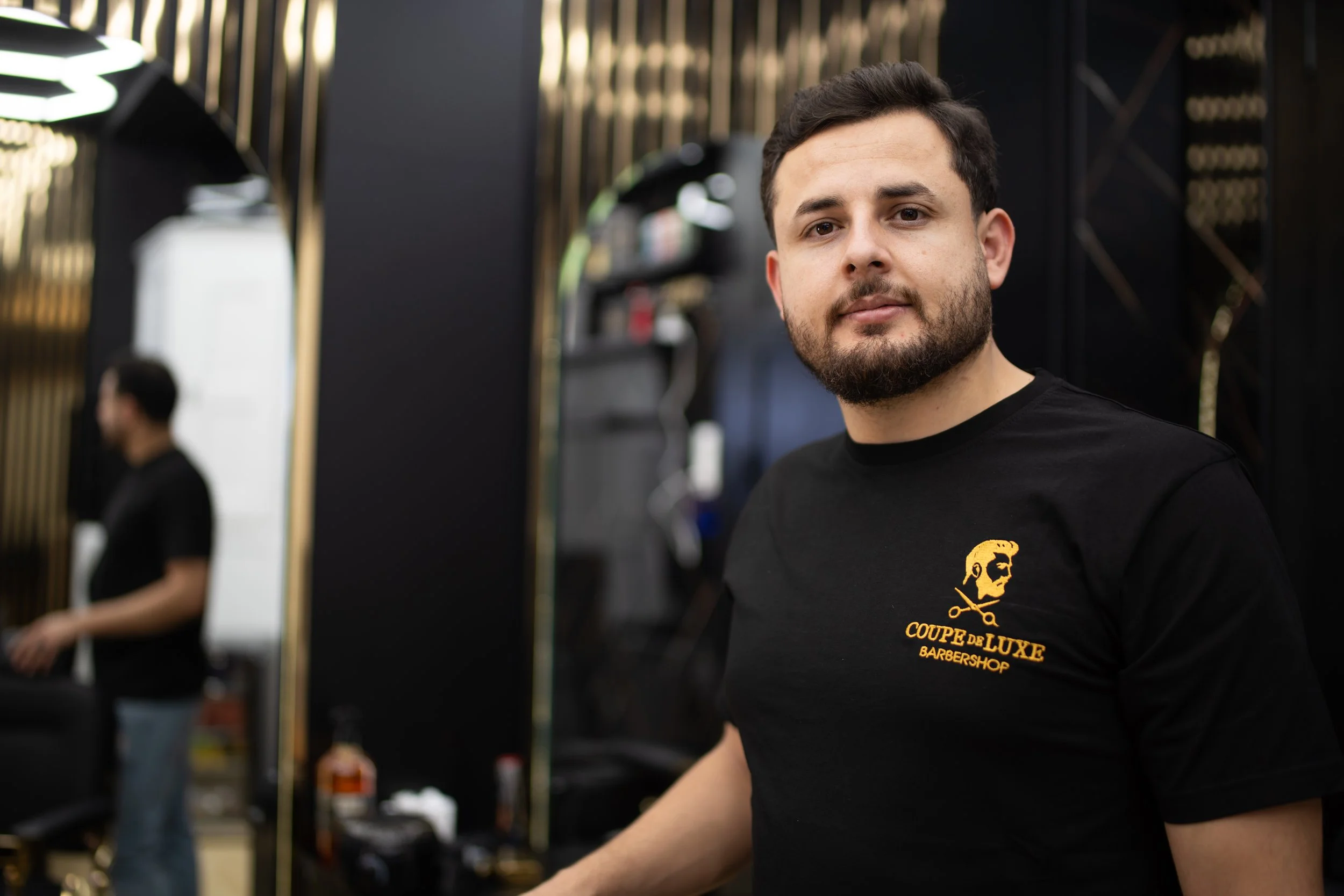 Friseur in einem Barbershop trägt schwarzes T-Shirt mit Logo. Blick in die Kamera, Hintergrund spiegeln und Friseur, der an einem Spiegel arbeitet.