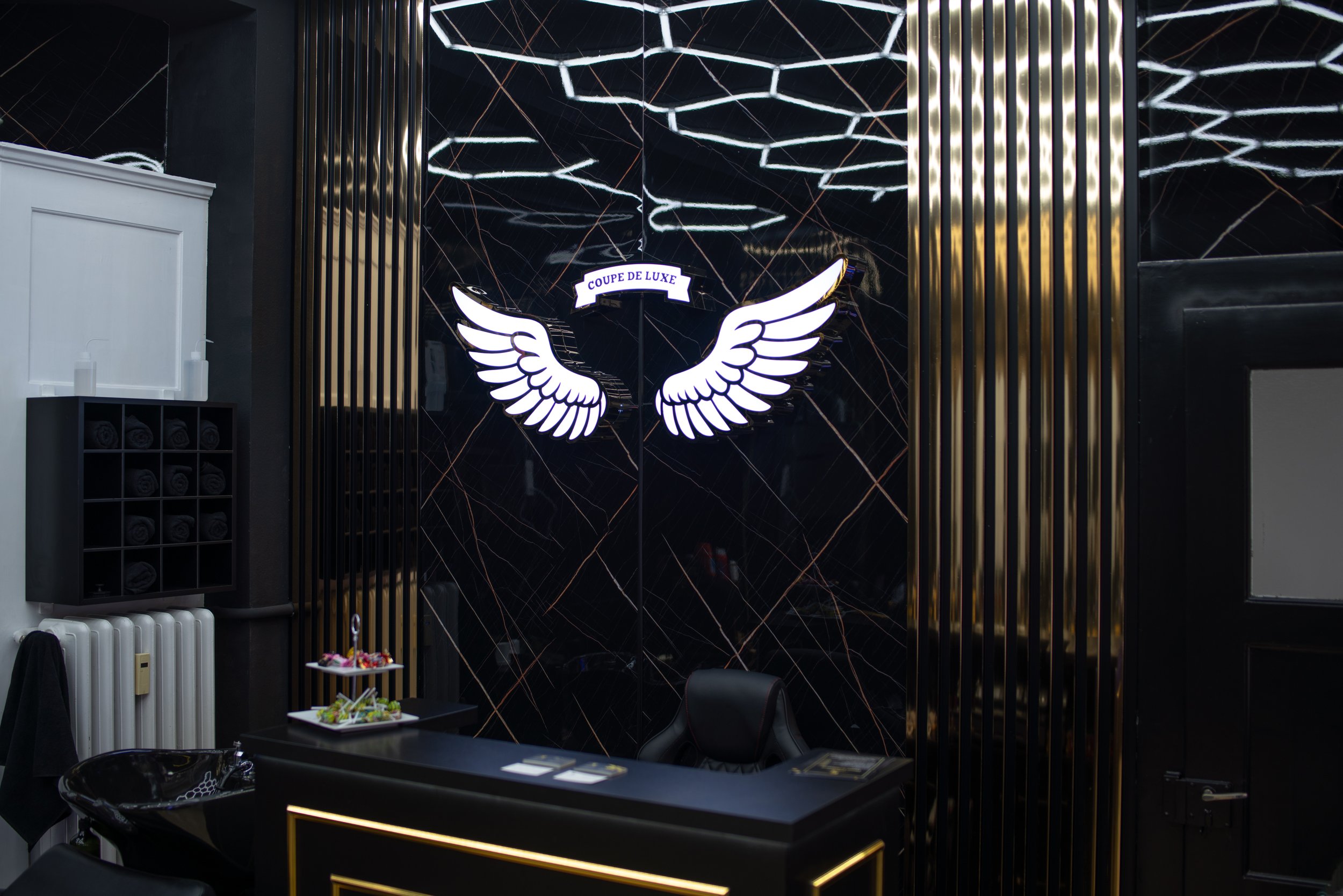 Empfangstresen mit schwarzem und goldenem Design, hinter dem eine Wand mit einem Lichtlogo mit Flügeln und der Aufschrift 'Coupe de Luxe' ist, in einem luxuriösen Raum mit dunklen Wänden und moderner Beleuchtung.