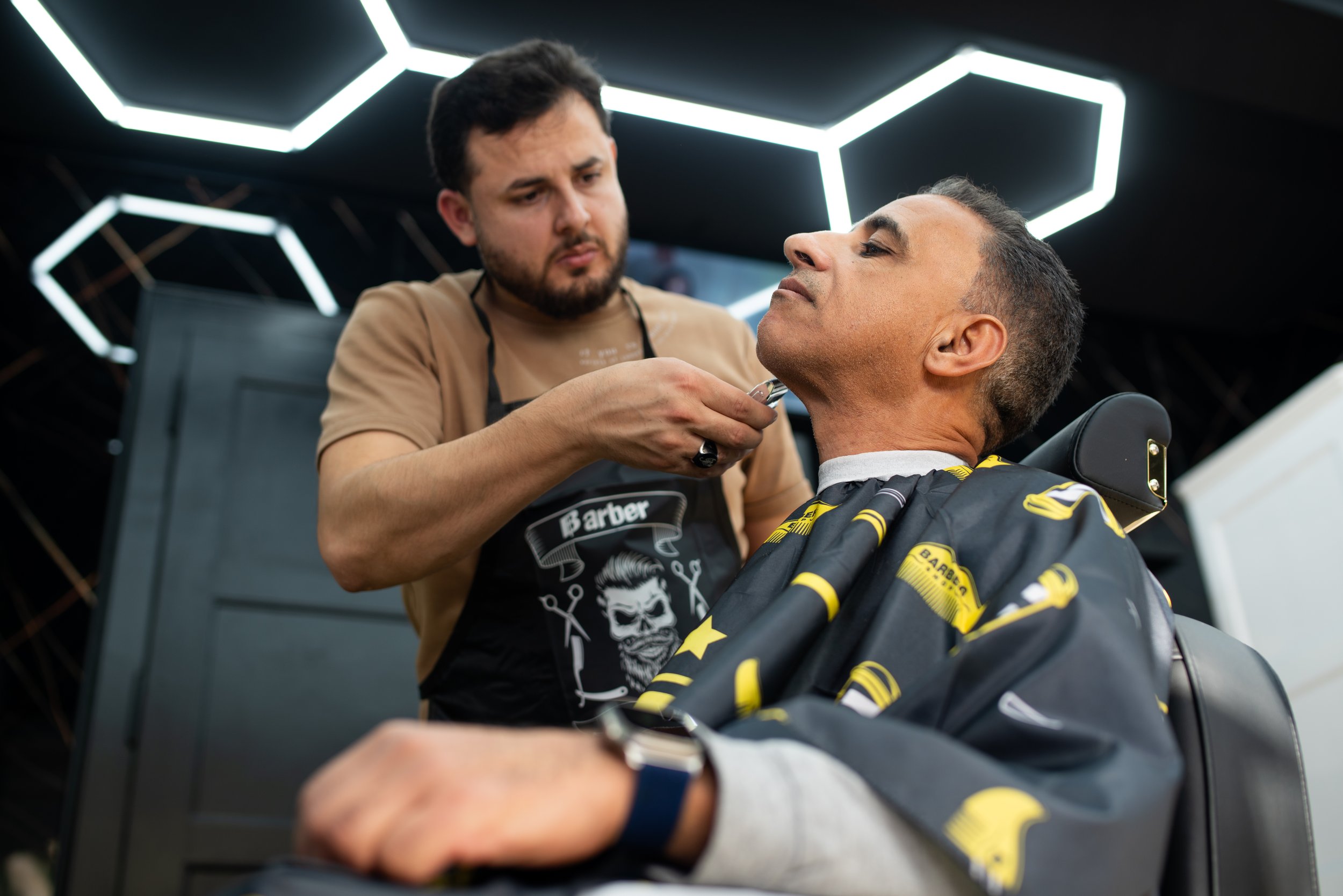 Friseur rasiert den Bart eines Mannes in einem Friseursalon mit moderner Beleuchtung an der Decke.