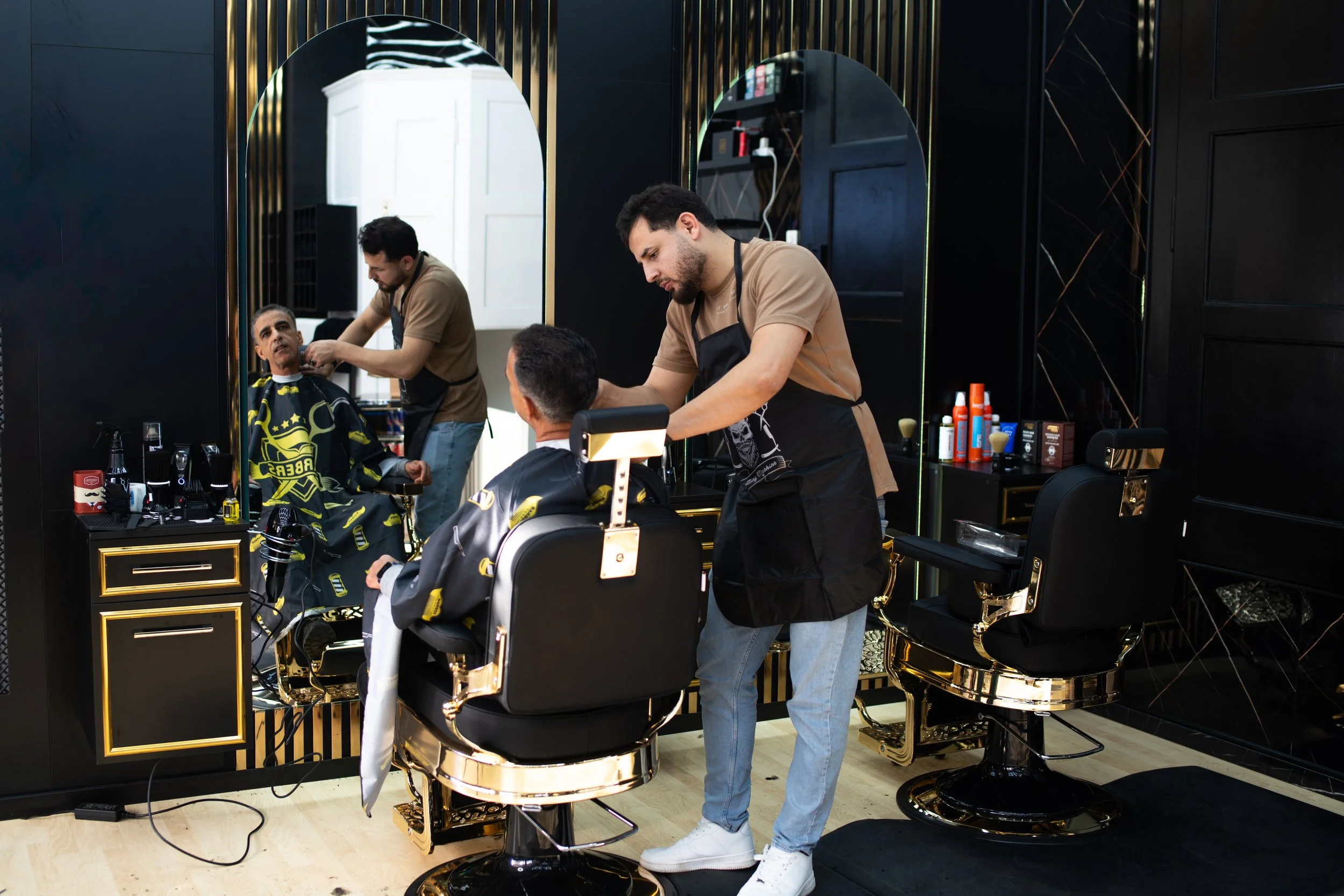 Ein Friseur schneidet einem Kunden die Haare in einem modernen Friseursalon mit schwarzen Wänden und goldverzierten Friseurstühlen.
