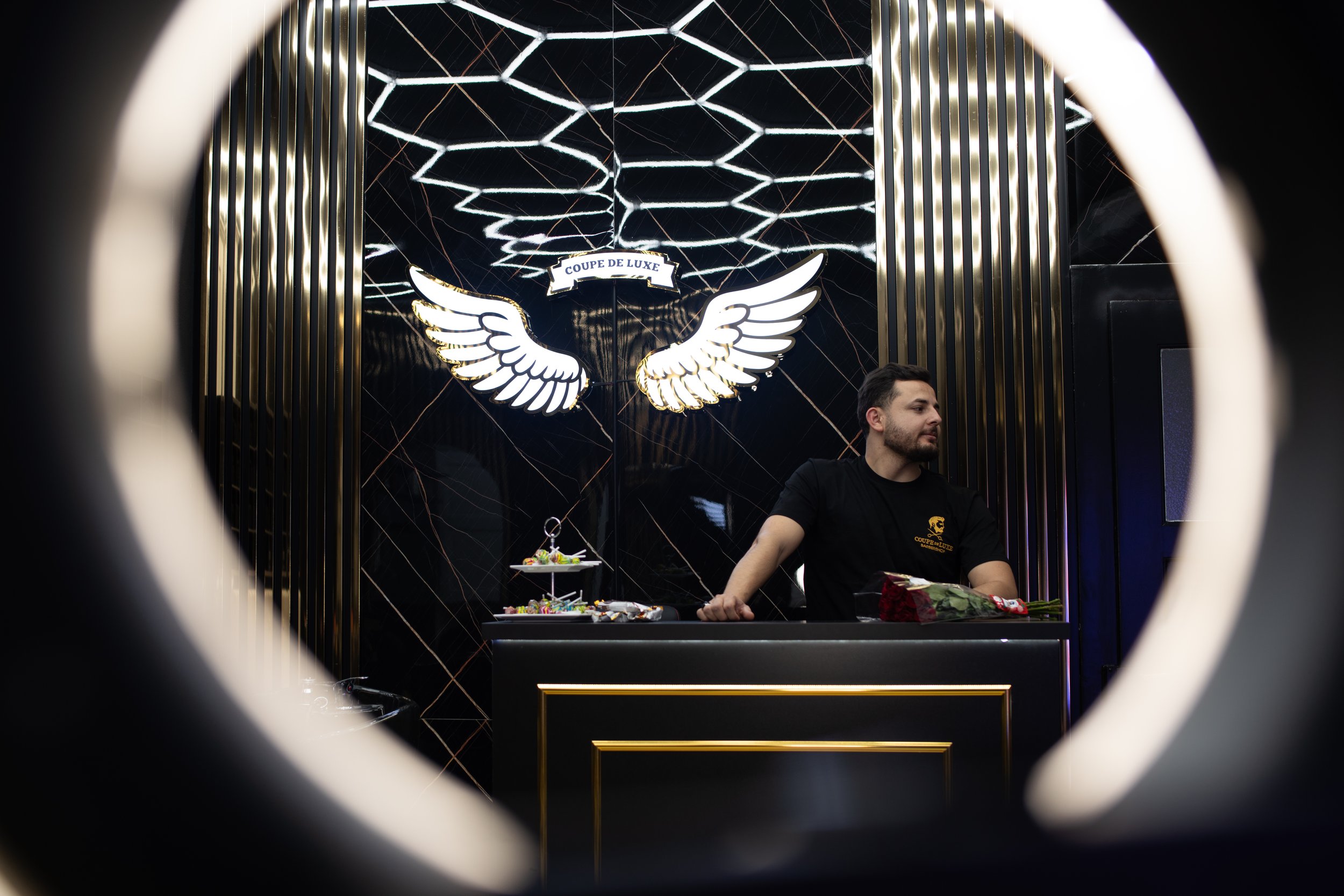 Ein Mann steht hinter einem schwarzen Empfangstresen in einem luxuriösen, dunklen Raum mit goldenen Akzenten. Im Hintergrund sind leuchtende Flügel aus Licht und ein Schild mit der Aufschrift "Coupe de Luxe" sichtbar.